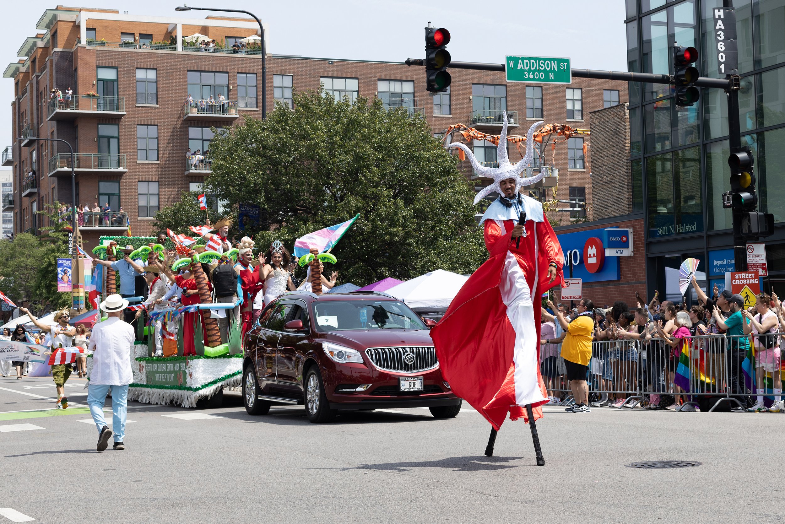 Parade with a float carrying people in colorful costumes, a person in a red and white costume with a headpiece and microphone, and spectators behind a barricade on a city street at the intersection of West Addison Street.