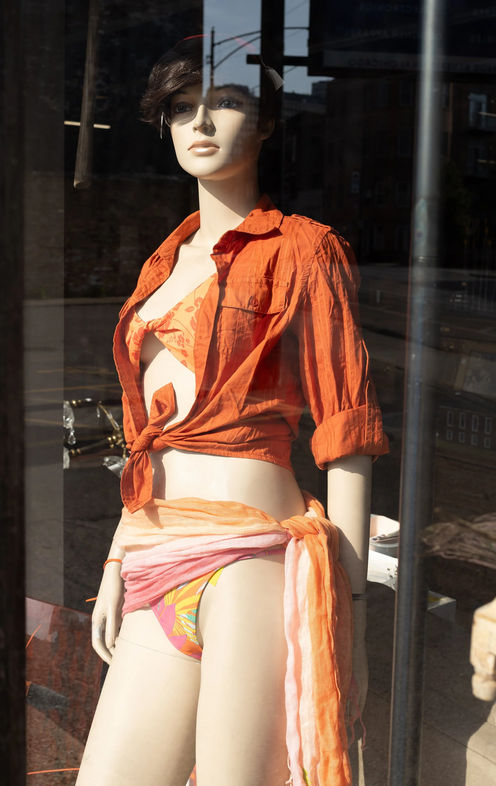 Mannequin dressed in an orange button-up shirt tied at the waist, with a colorful bikini bottom and a lightweight scarf around the hips, displayed outside a store window.