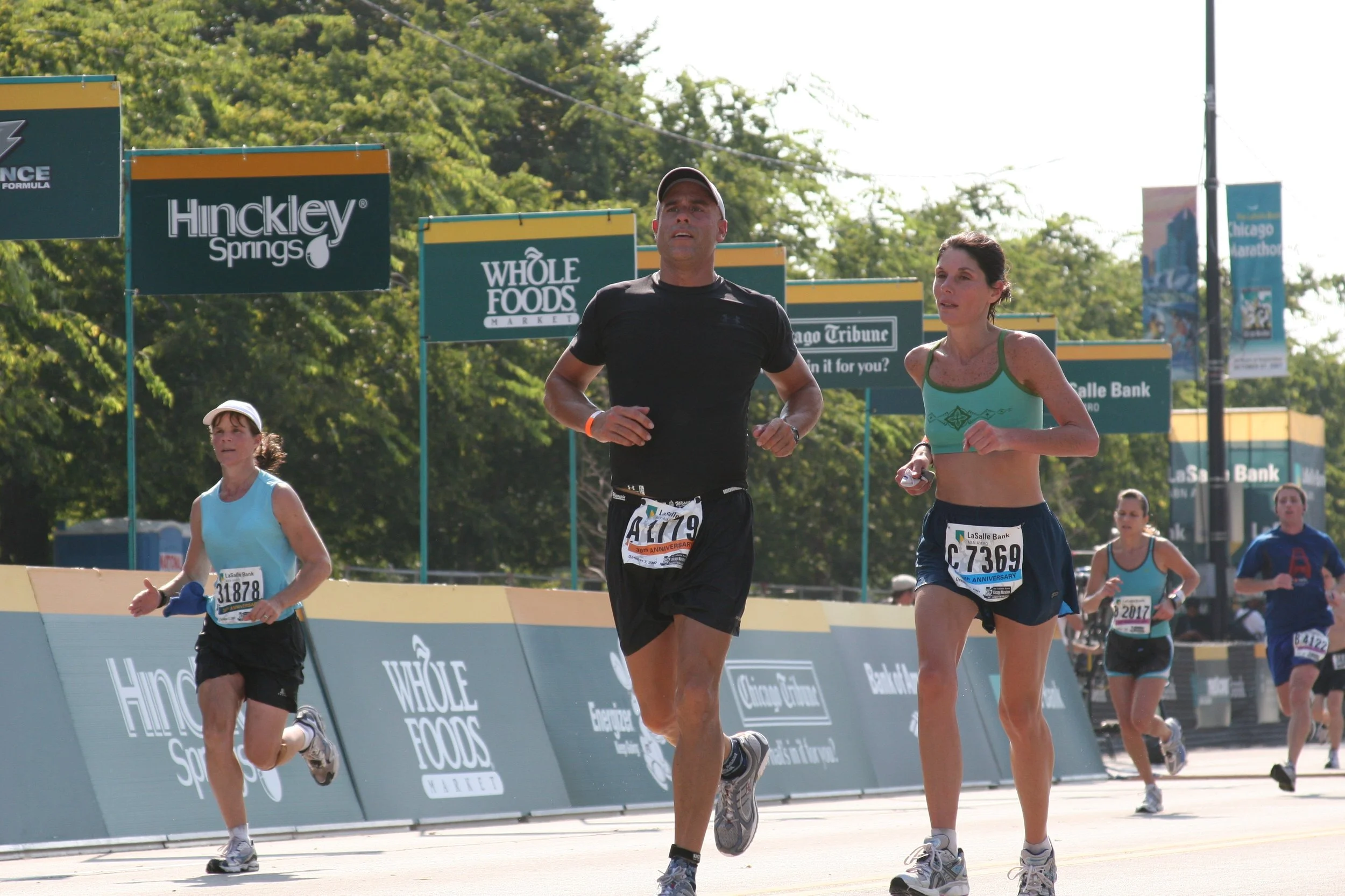 Several runners participating in a race, with banners and advertisements in the background, some of which read 'Hinckley Springs', 'Whole Foods', and 'Chicago Tribune'.