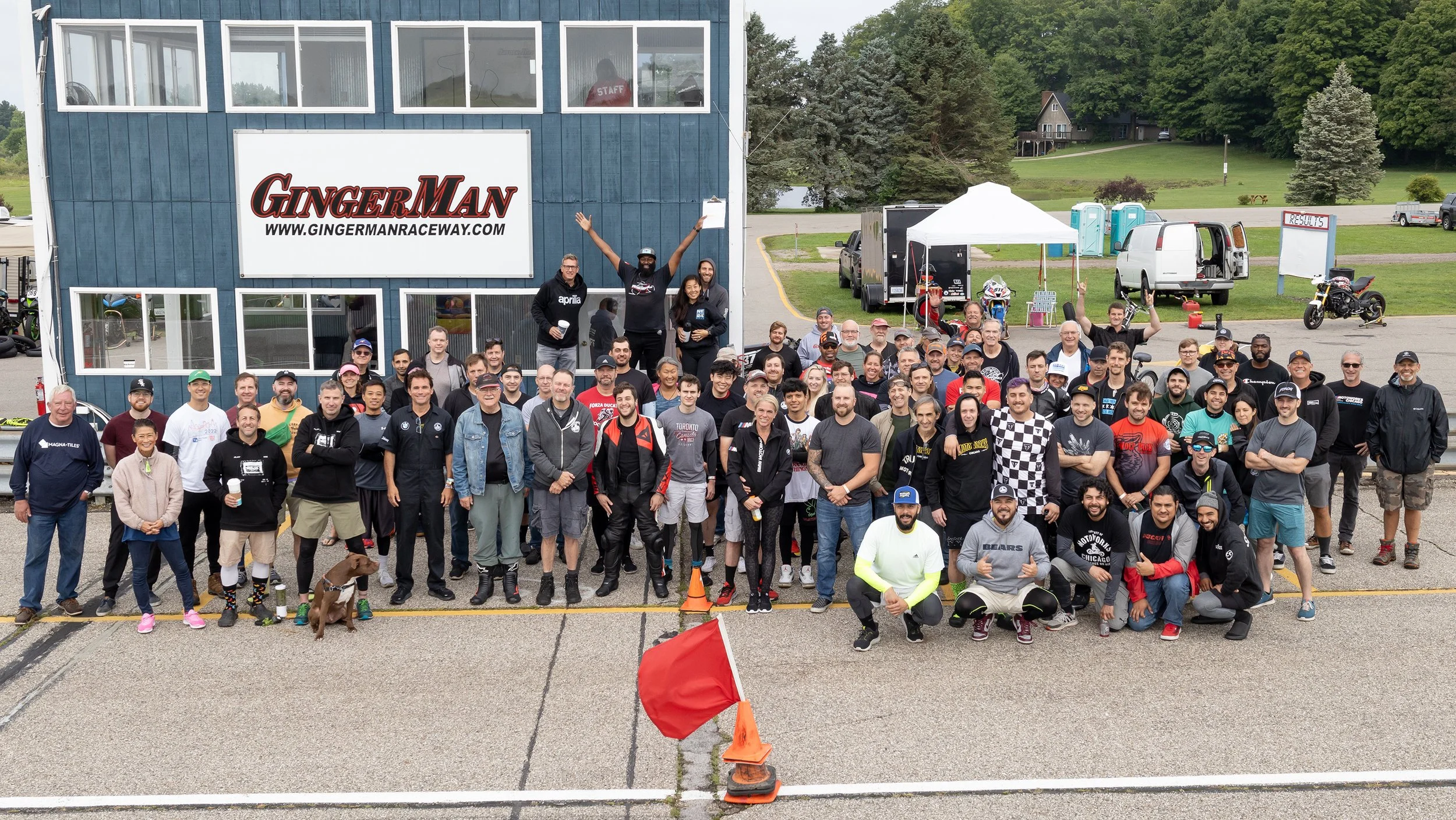 Large group of diverse people gathered at a raceway, posing for a group photo in front of a sign that reads 'GingerMan Raceway'. The group includes men, women, and children, some holding drinks, with a few raising their hands. The setting appears to 