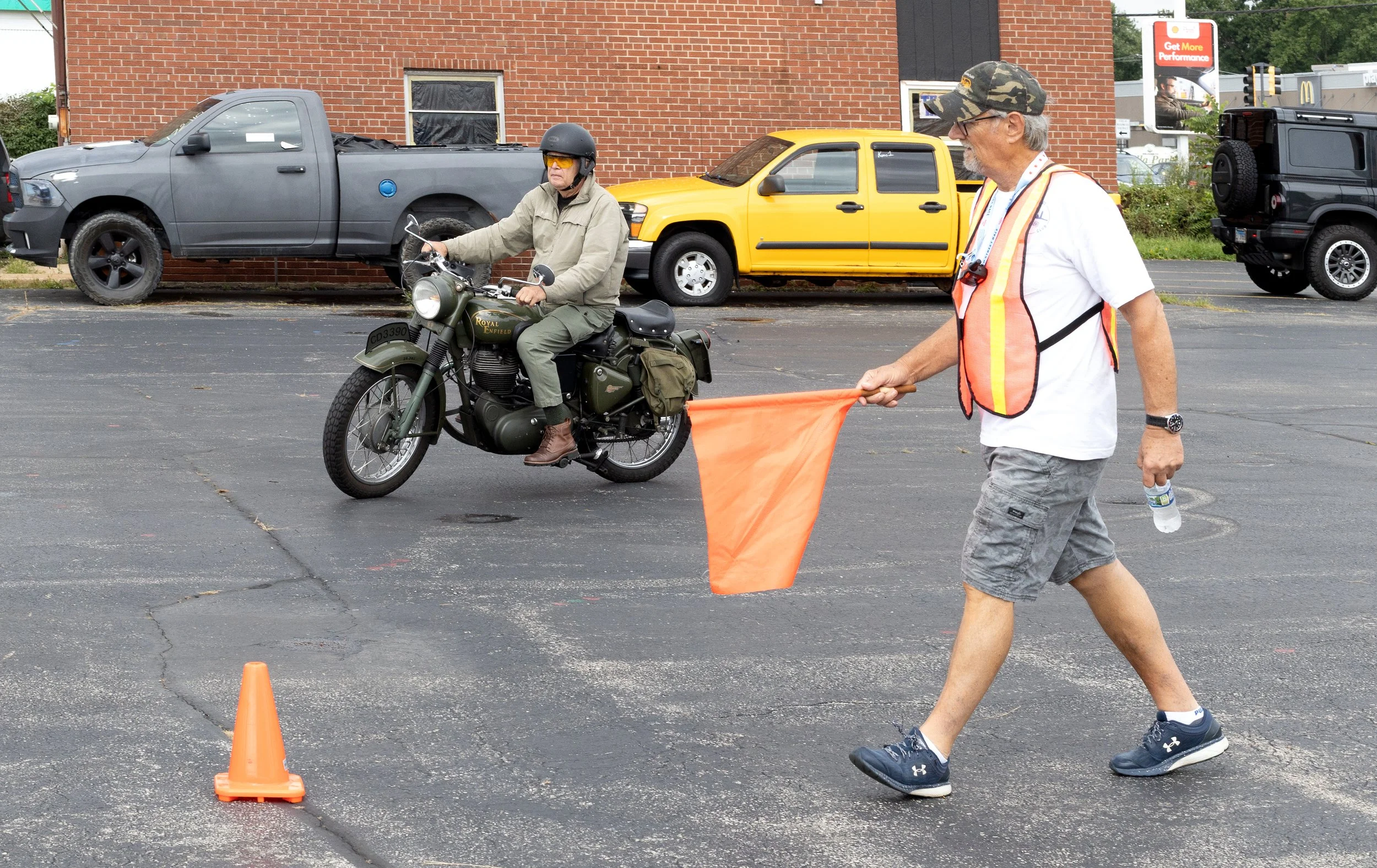 A man in a white t-shirt, camouflage hat, glasses, and gray shorts holding an orange flag walks past a man riding a motorcycle, wearing a helmet and sunglasses, on a paved lot with orange cones and parked cars in the background.