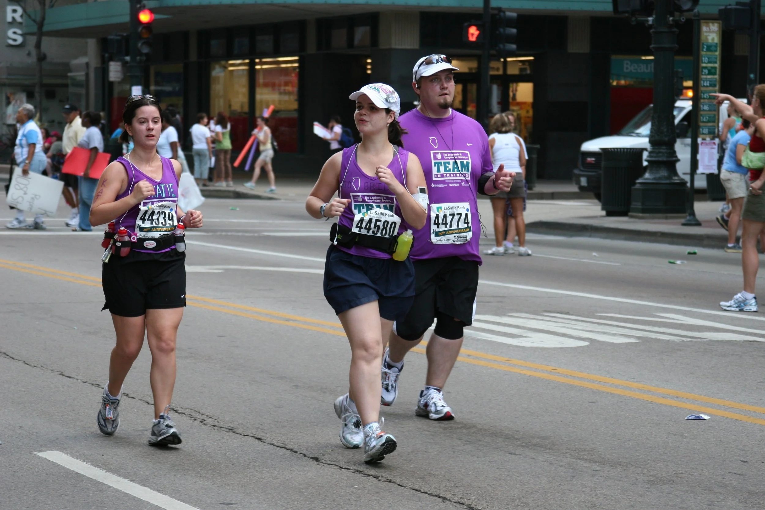 Three runners participating in a road race, wearing purple shirts and bib numbers, crossing an intersection with spectators in the background.