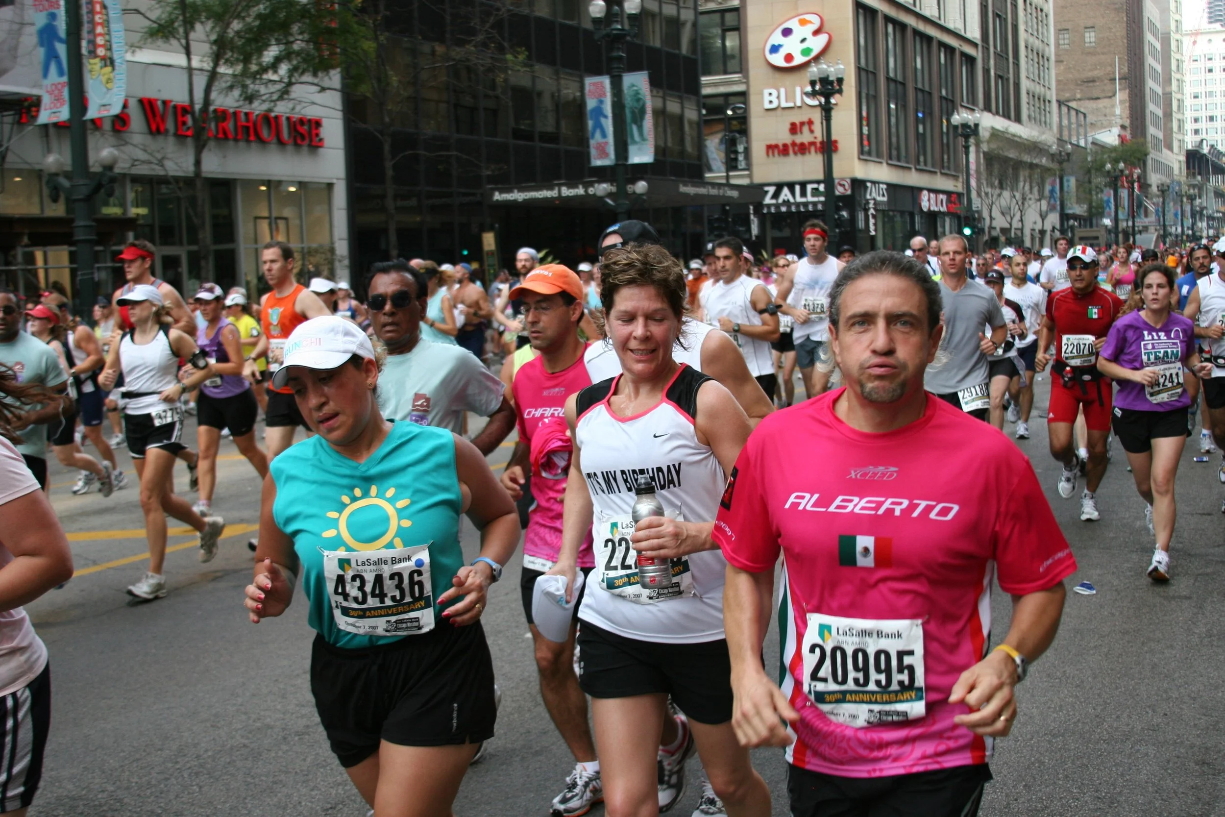 A large group of marathon runners participating in a race on a city street. The runners are wearing athletic clothing, bibs with numbers, and some are holding water bottles. The background shows tall city buildings with signs, including a jewelry sto