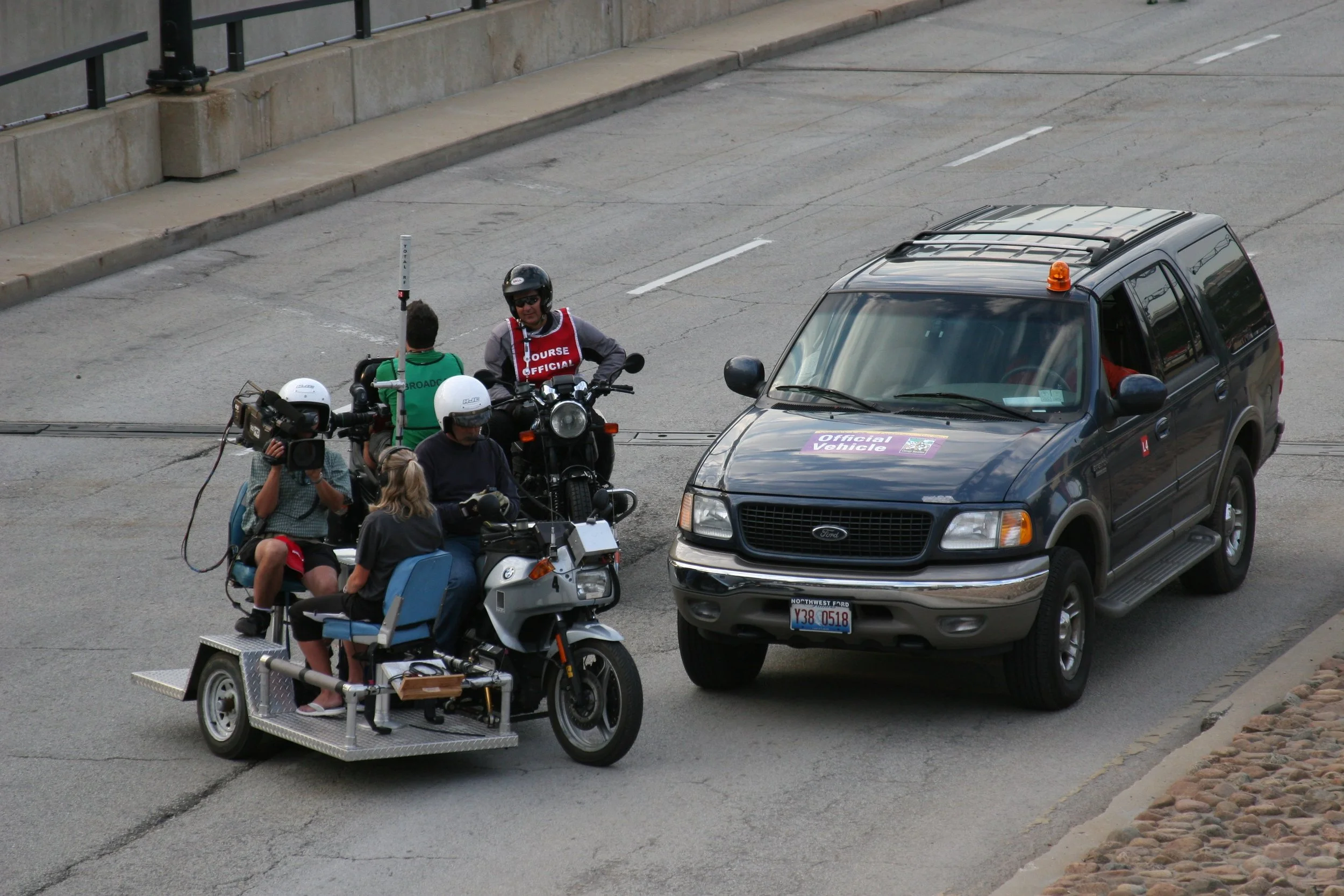 A group of people riding motorcycles and scooters on a city street, with one person on a three-wheeled vehicle and a black SUV with an orange light on top nearby.