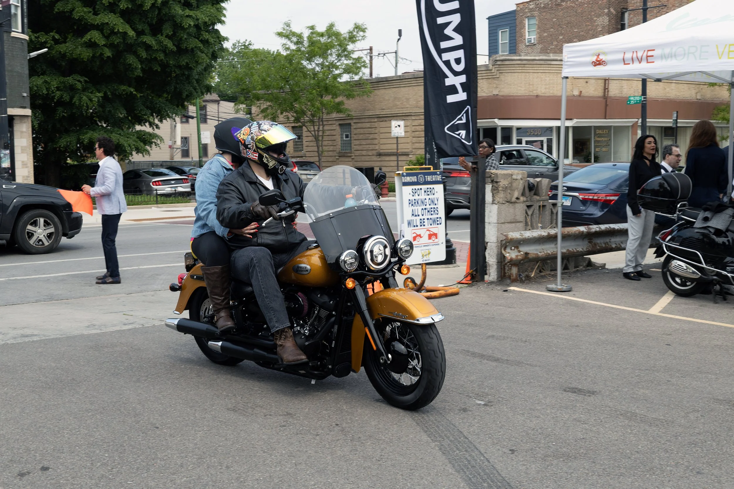 Two motorcyclists wearing helmets riding a brown Harley Davidson motorcycle on a city street. In the background, pedestrians and parked cars are visible, with storefronts and trees lining the street.