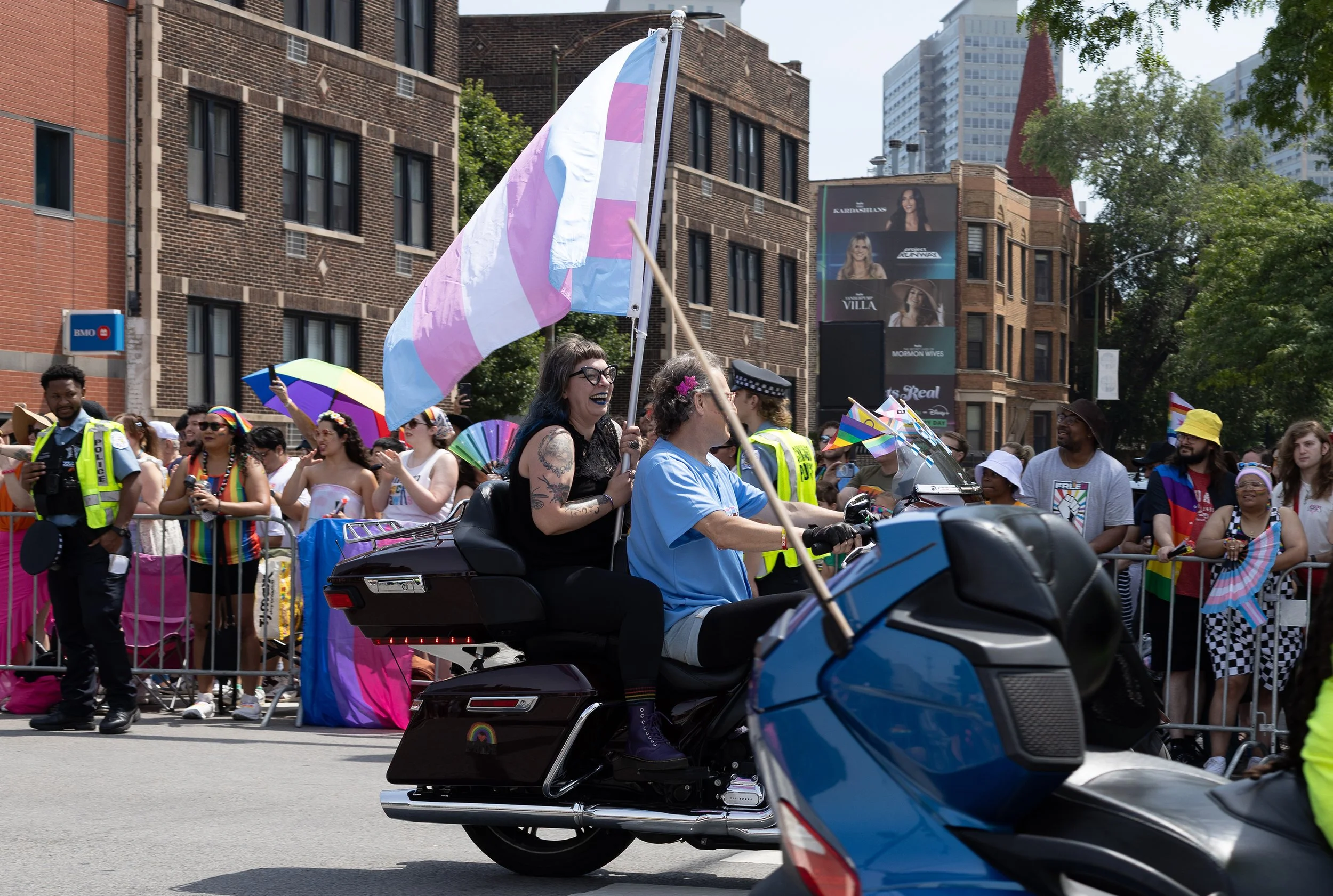 Group of people at a pride parade, some holding rainbow flags, some wearing rainbow accessories, and a woman riding a motorcycle with others in the background.