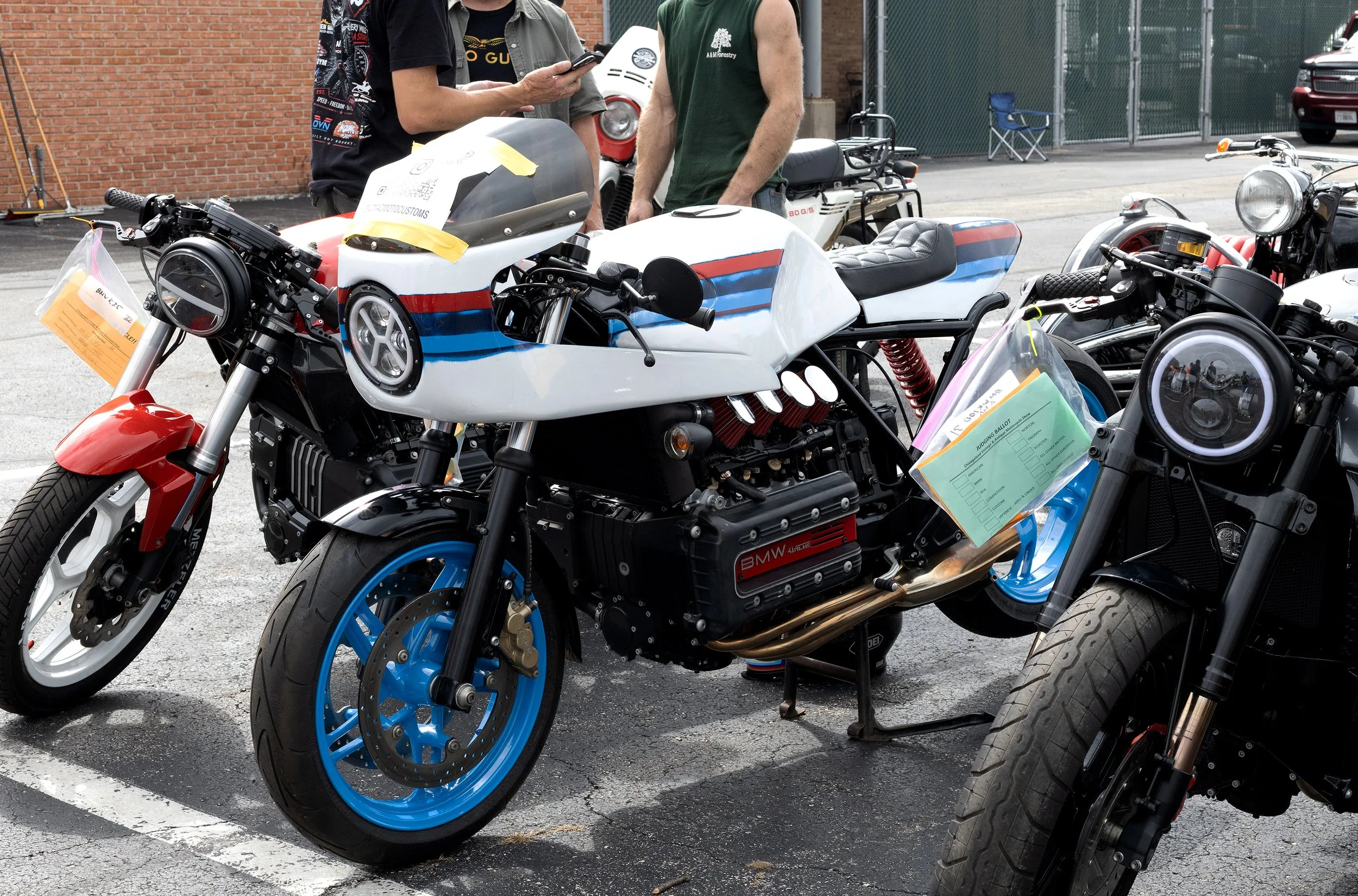 Several custom motorcycles on display outdoors, with people examining them. The bikes have unique paint jobs and modifications, and some have information tags attached.