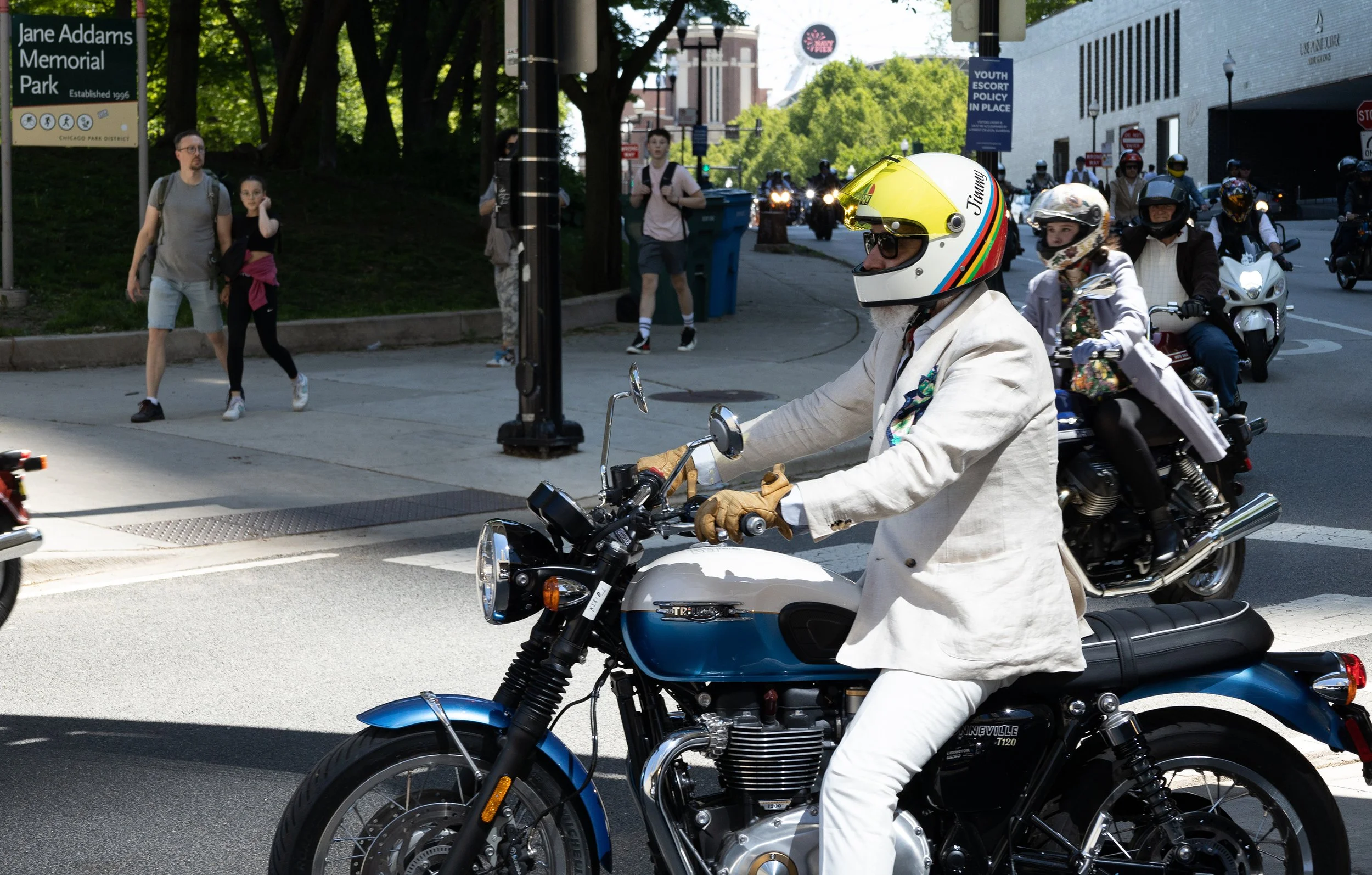 A man in a cream-colored suit and a colorful helmet riding a blue Triumph motorcycle on a city street. Several pedestrians walking on the sidewalk and more motorcyclists riding behind him are visible, along with street signs and trees.