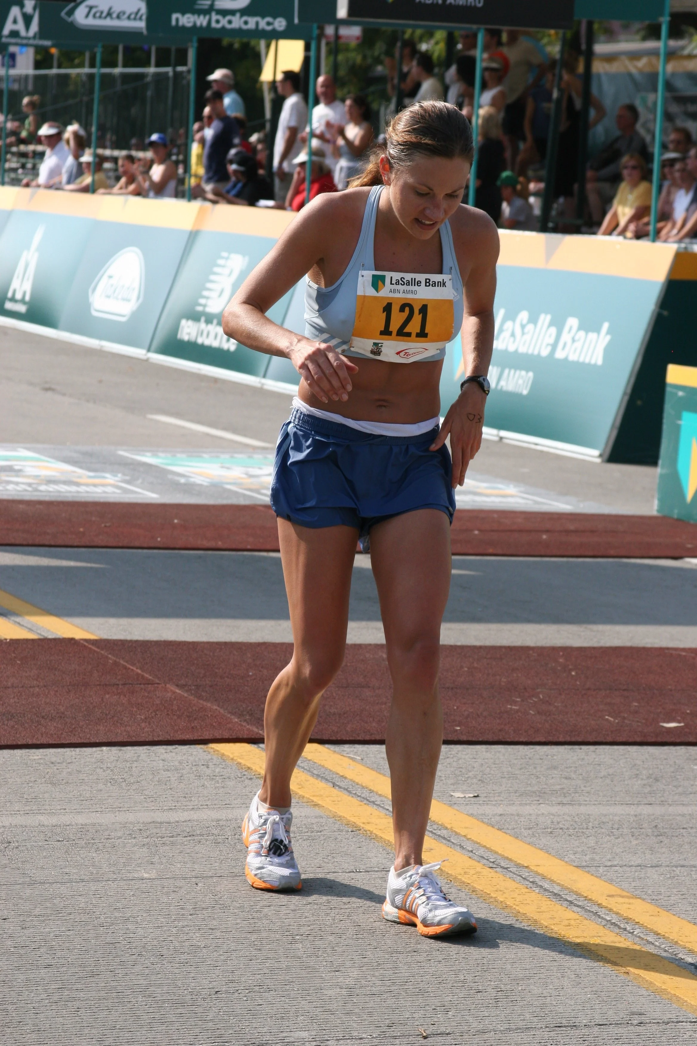 A female athlete wearing a light blue tank top, blue shorts, and running shoes crosses the finish line of a race, with a yellow number 121 bib pinned to her top, and spectators watching from the stands behind a barricade.