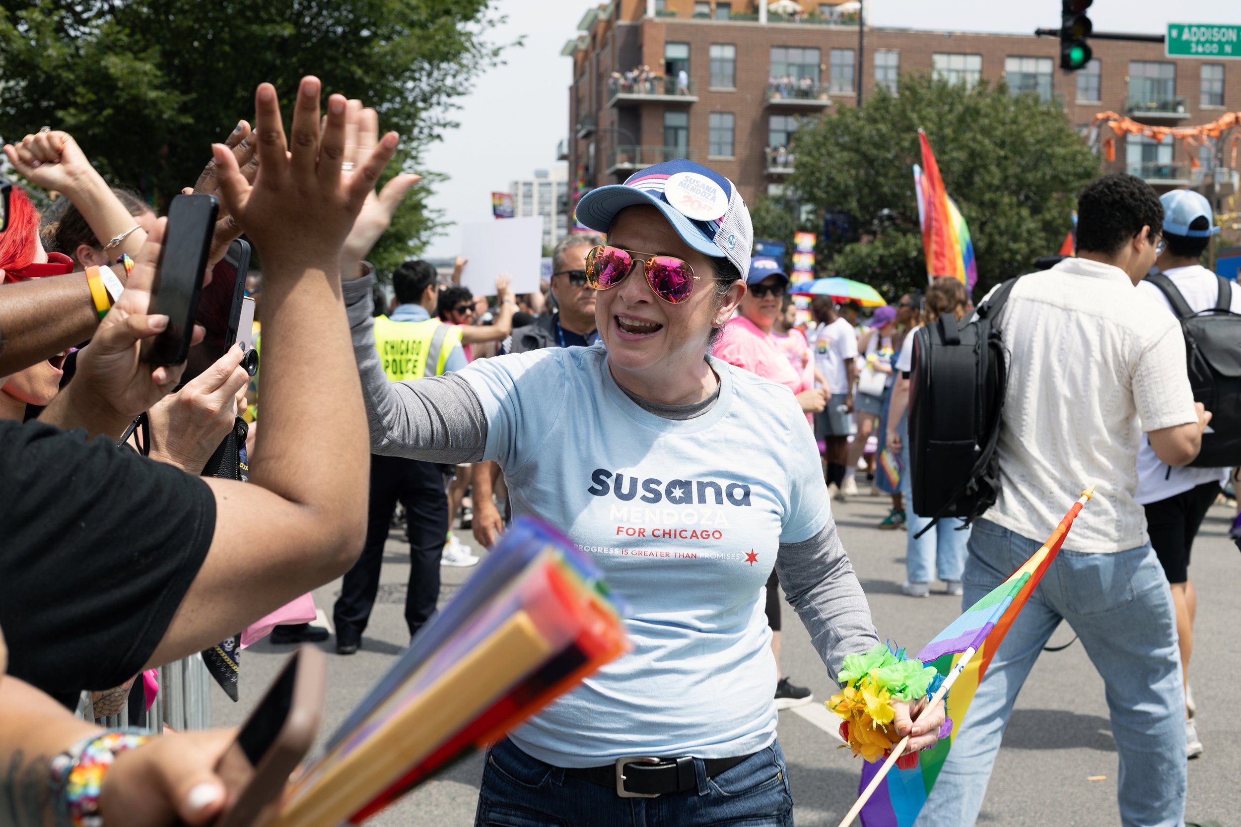 A woman in sunglasses and a campaign t-shirt high-fives a person at a pride parade. She wears a cap with campaign buttons and is holding rainbow flags and rainbow-colored pom-poms. The street is filled with people, some holding rainbow flags, with ap