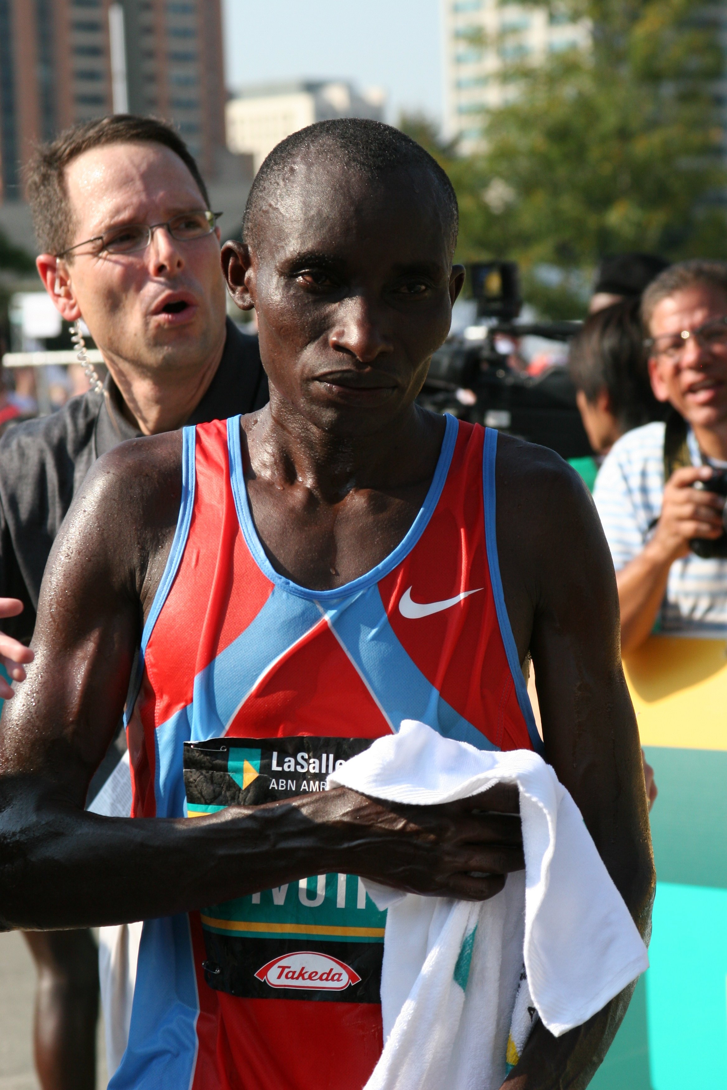 A male marathon runner in a colorful sleeveless running shirt holding a white towel after finishing a race, with a man and woman in the background celebrating.