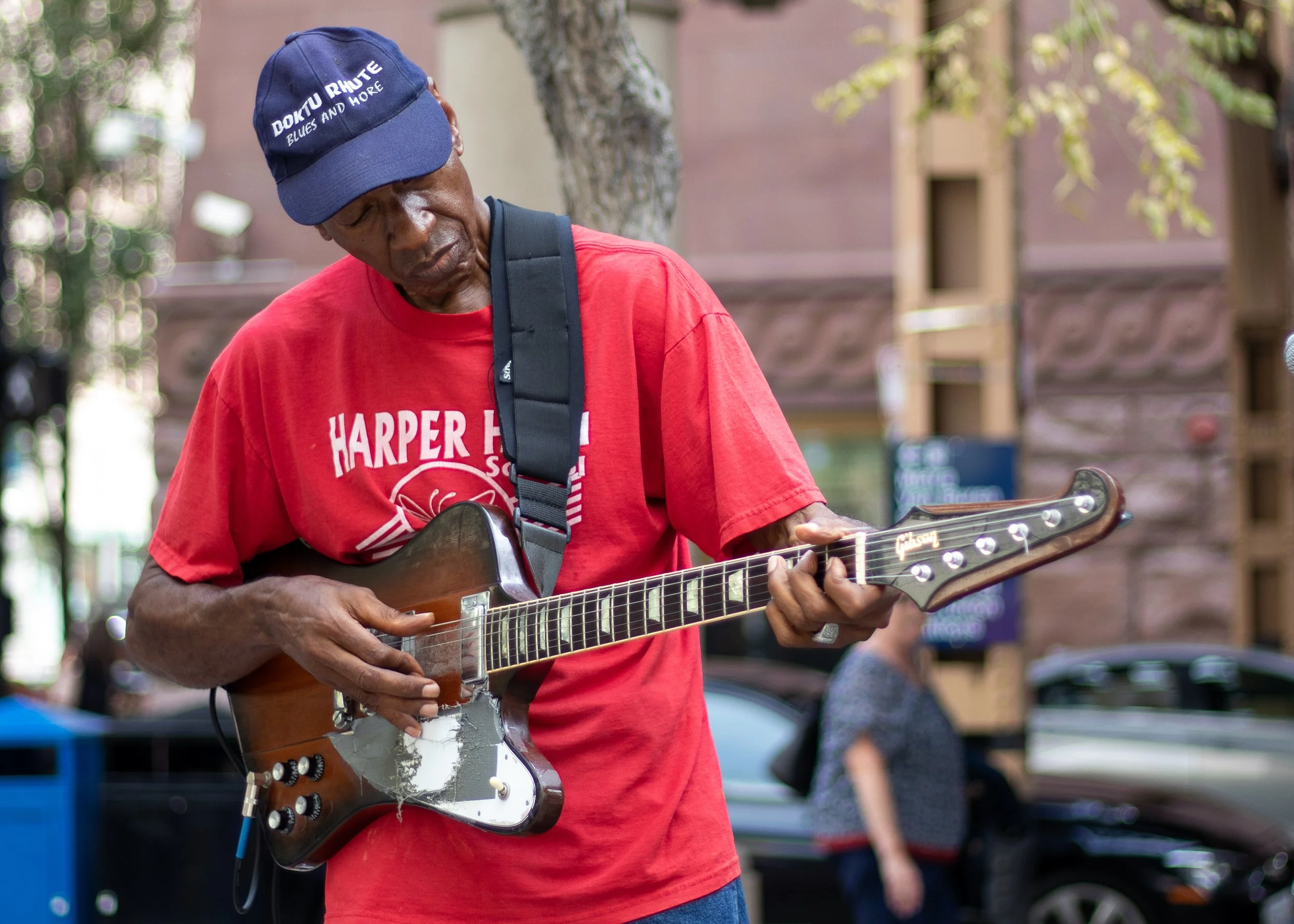 A man playing an electric guitar outdoors with trees and blurred background.