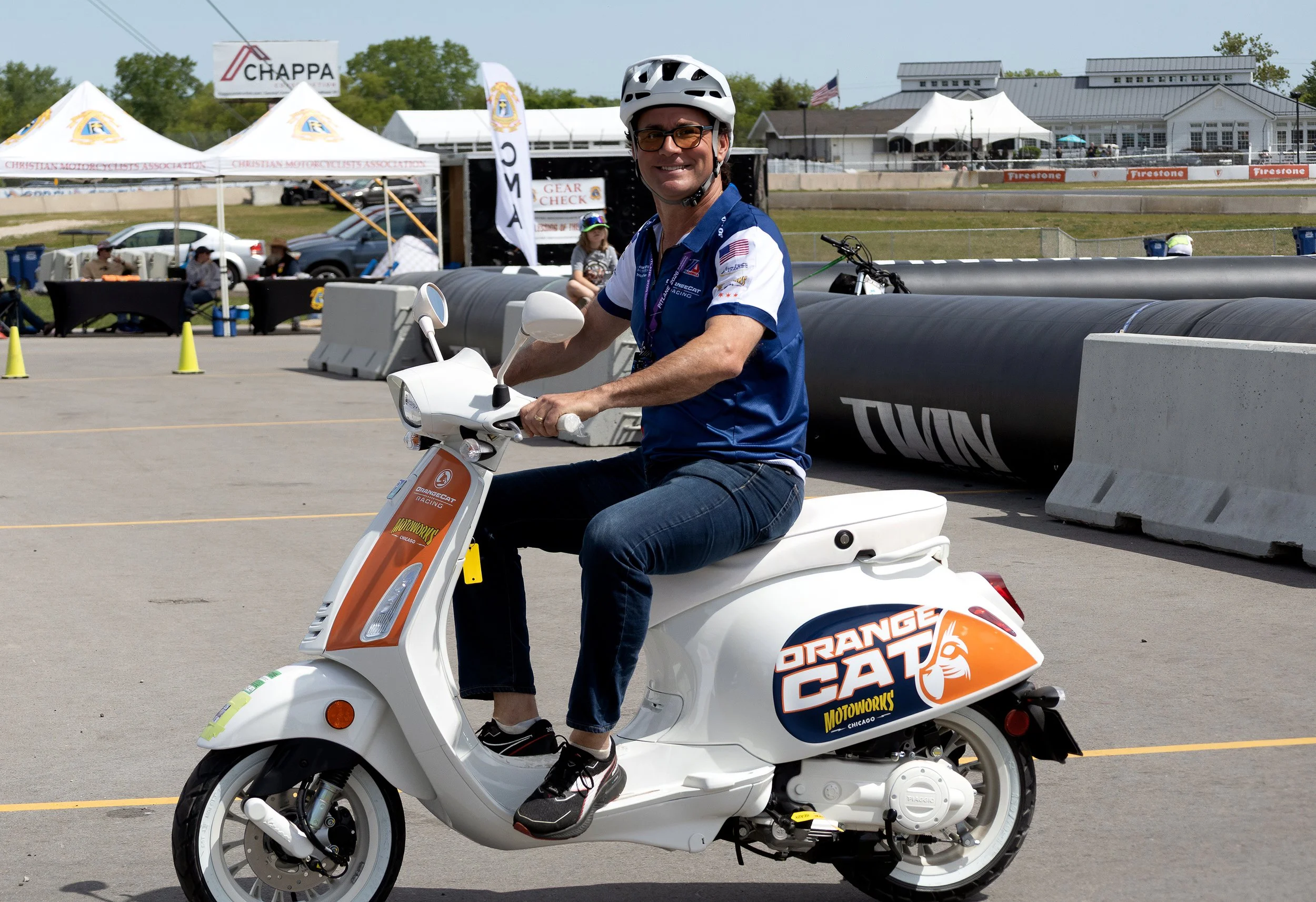 A man wearing a white helmet, glasses, and a racing shirt is riding a white scooter with orange and black graphics at an outdoor event with tents and people in the background.