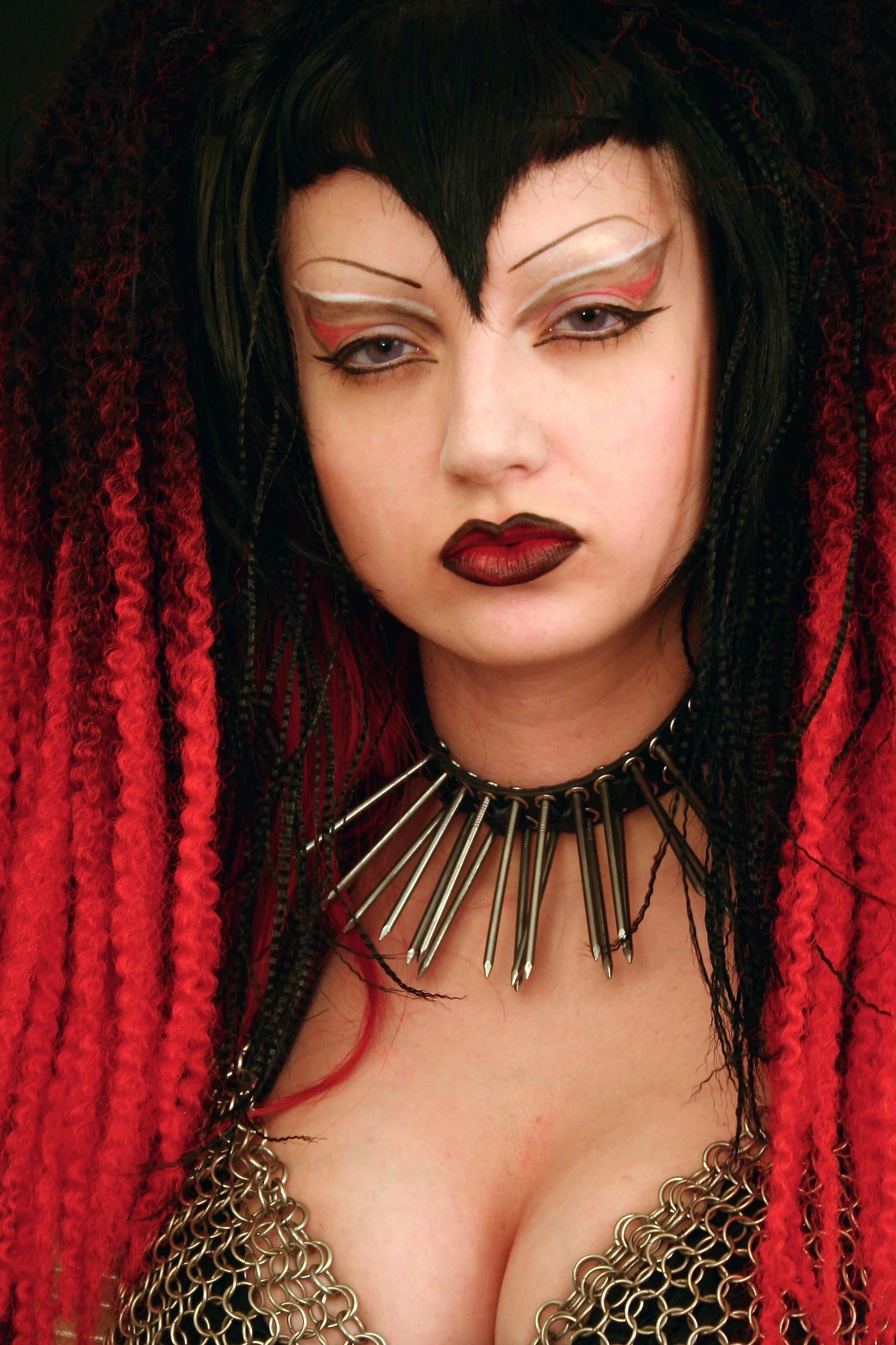 A person with pale skin, dark makeup, and elaborate black and red hair styled in dreadlocks. They are wearing metallic chain jewelry and a spiked necklace.