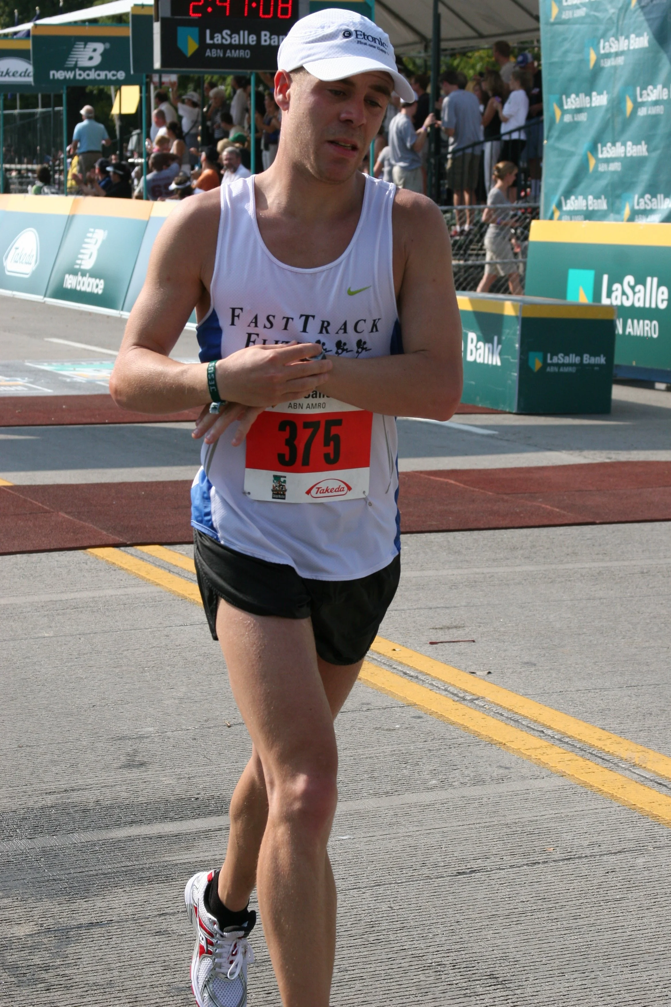 A male marathon runner with a bib number 375, wearing a white sleeveless shirt and a white cap, running on a marked street near the finish line of a race, with a digital timer and spectators in the background.