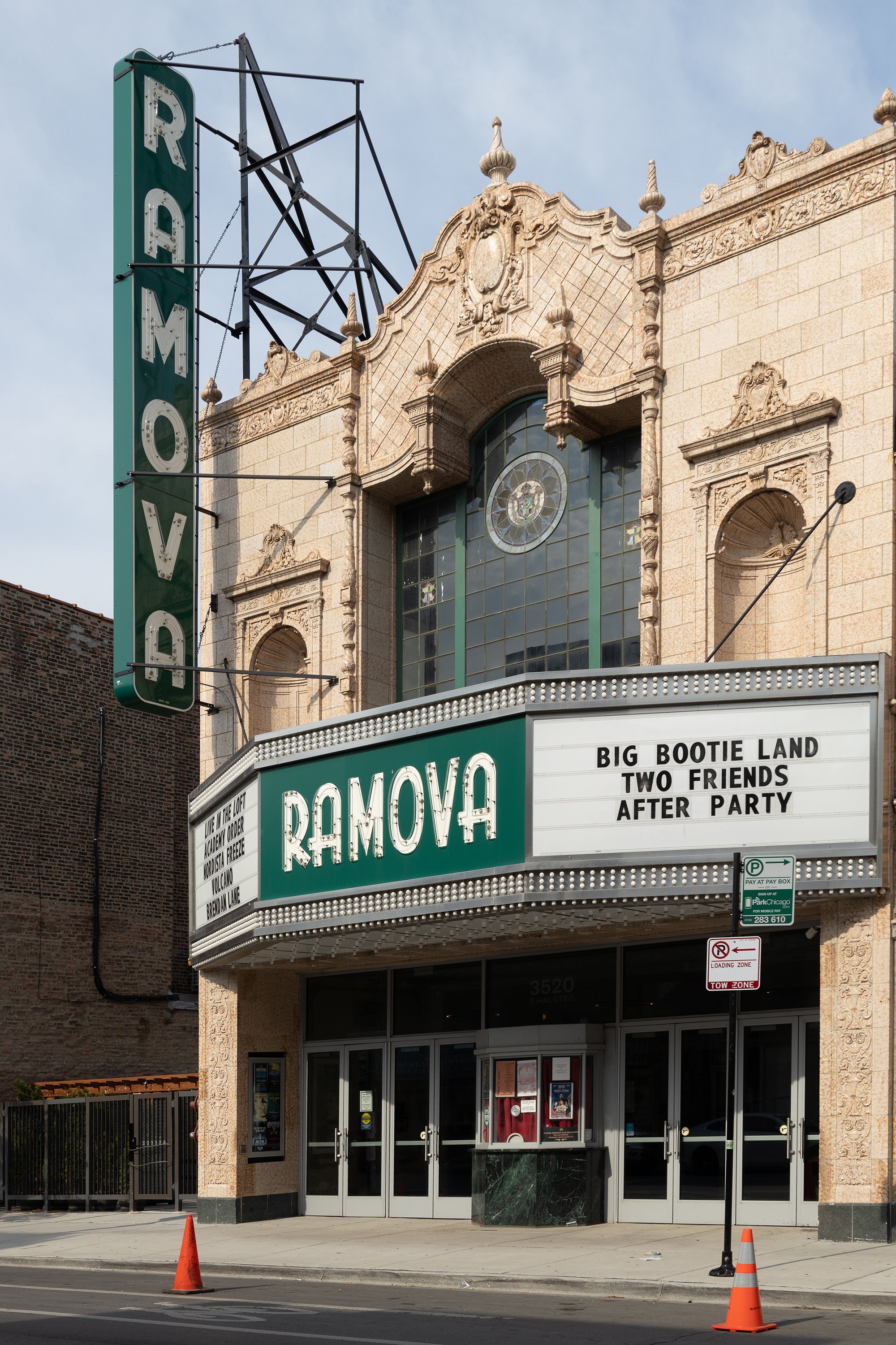 The facade of a movie theater named Ramova with a large marquee sign displaying song titles, and a vertical green sign with the name Ramova, alongside some traffic cones and parking signs outside.