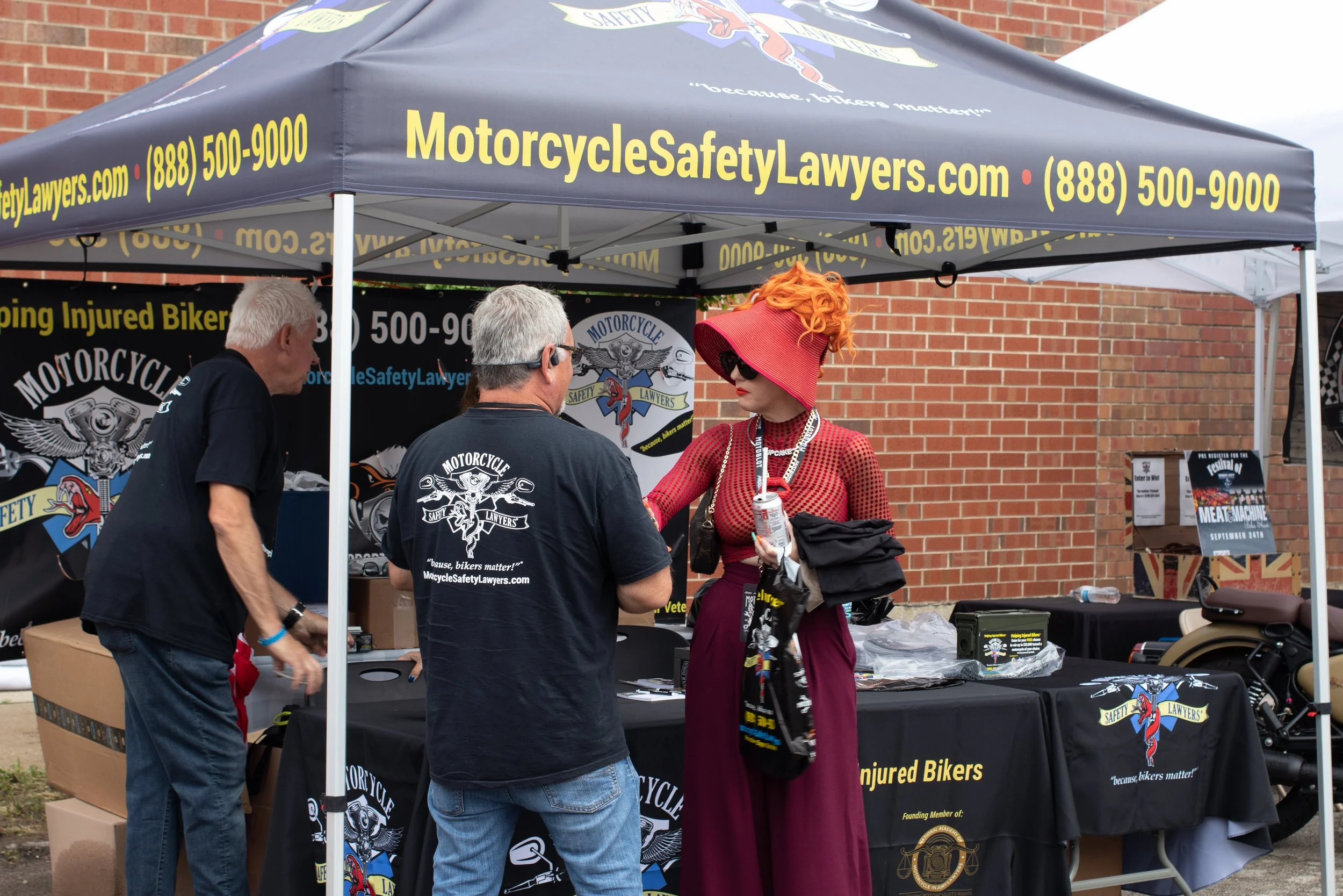 People at an outdoor booth for Motorcycle Safety Lawyers, with a banner displaying the website and phone number, and a woman with red hair, sunglasses, and a red hat talking to two men wearing black shirts with the logo.
