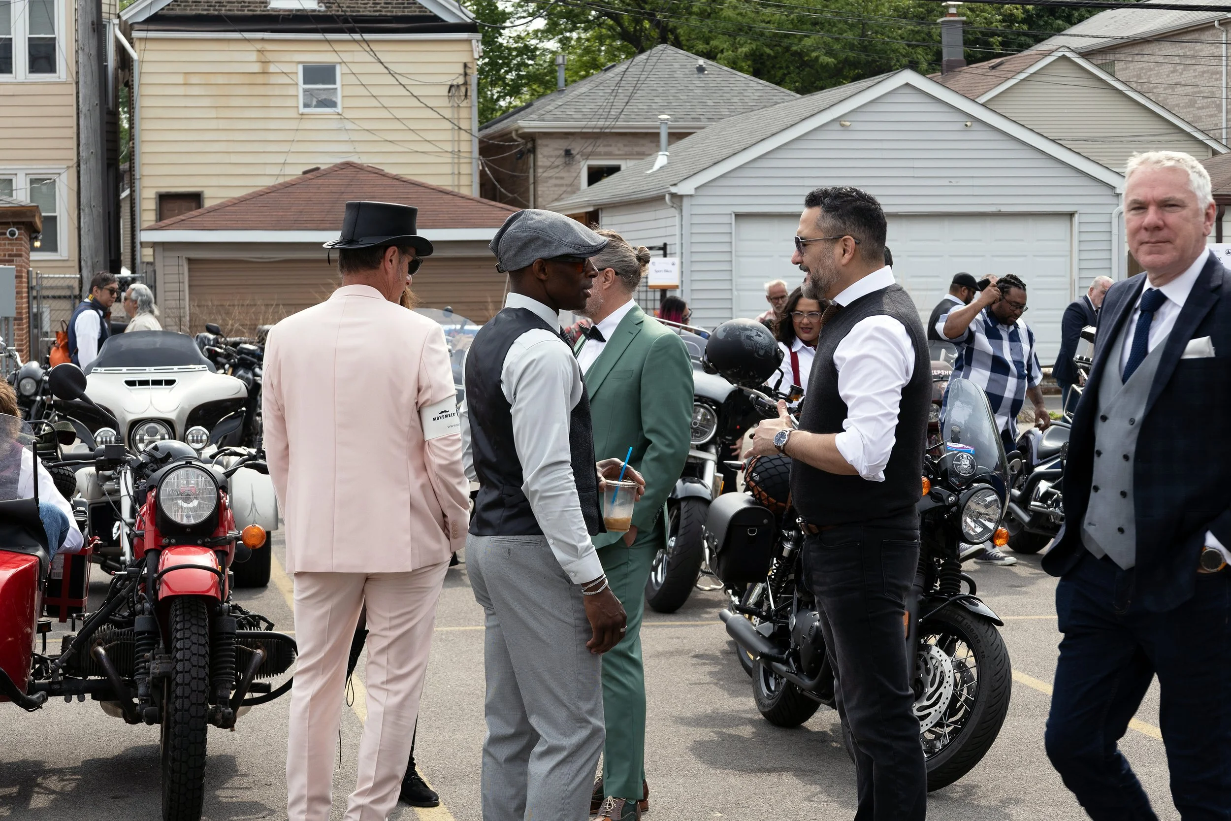 People gathered in an outdoor parking lot during a motorcycle event, talking and socializing among motorcycles and scooters, with houses in the background.