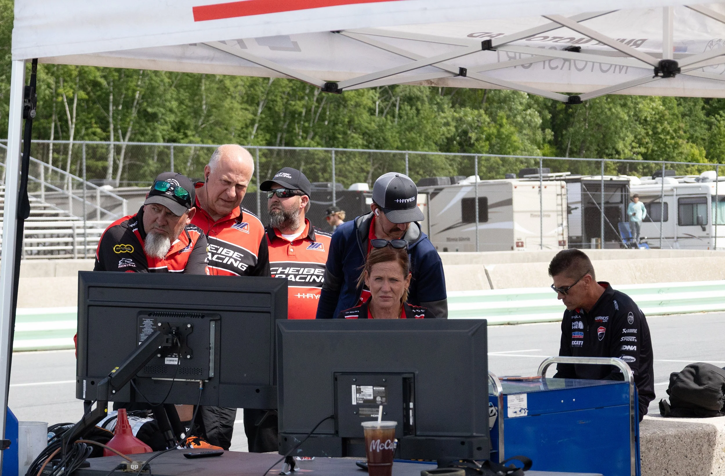 Group of people gathered under a white tent at a racetrack, looking at computer monitors, with RVs and trees in the background.