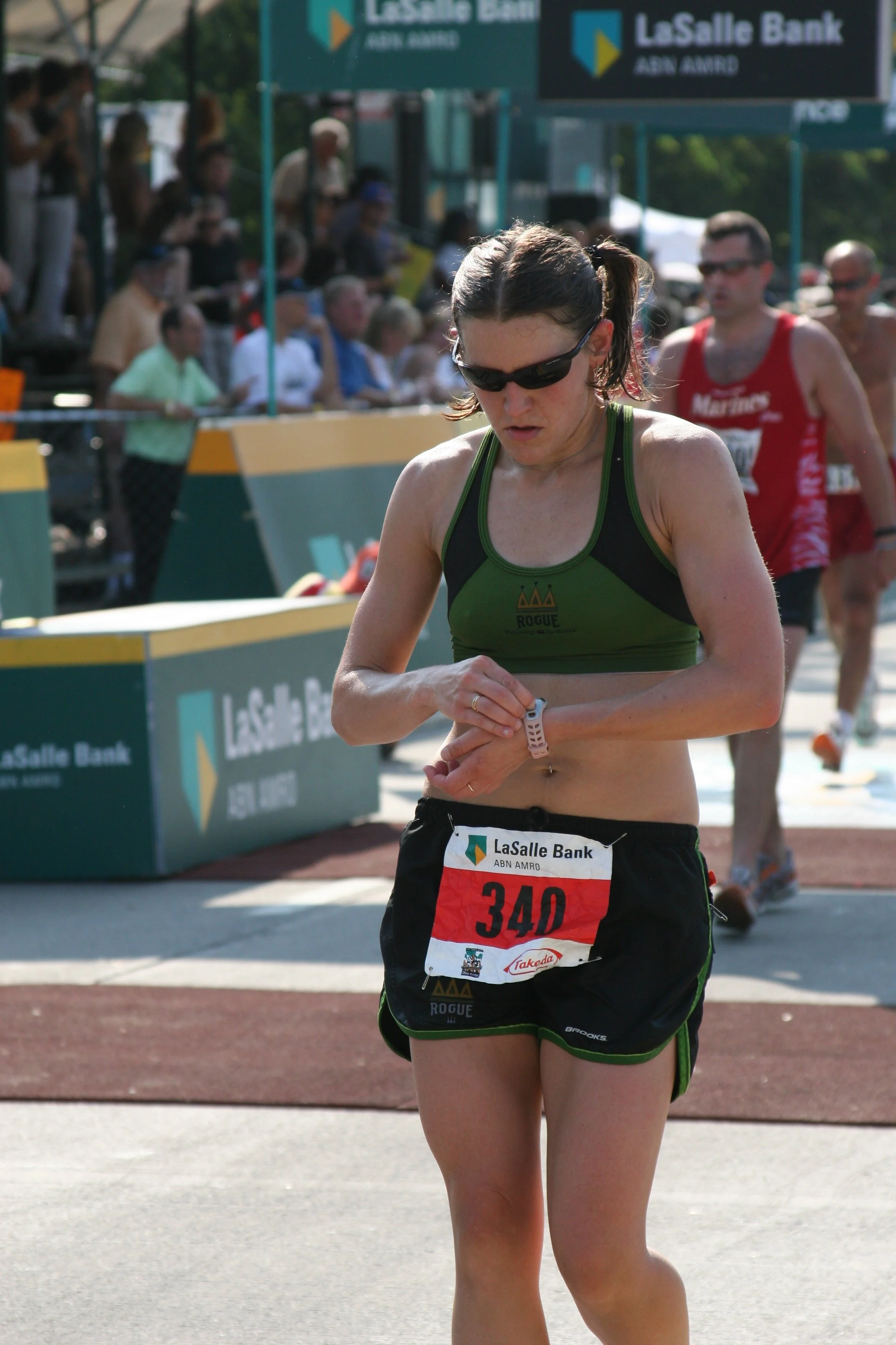 Female marathon runner checking her watch during a race, wearing a green and black athletic outfit, glasses, and a numbered bib. In the background, spectators observe and banners display 'LaSalle Bank'.