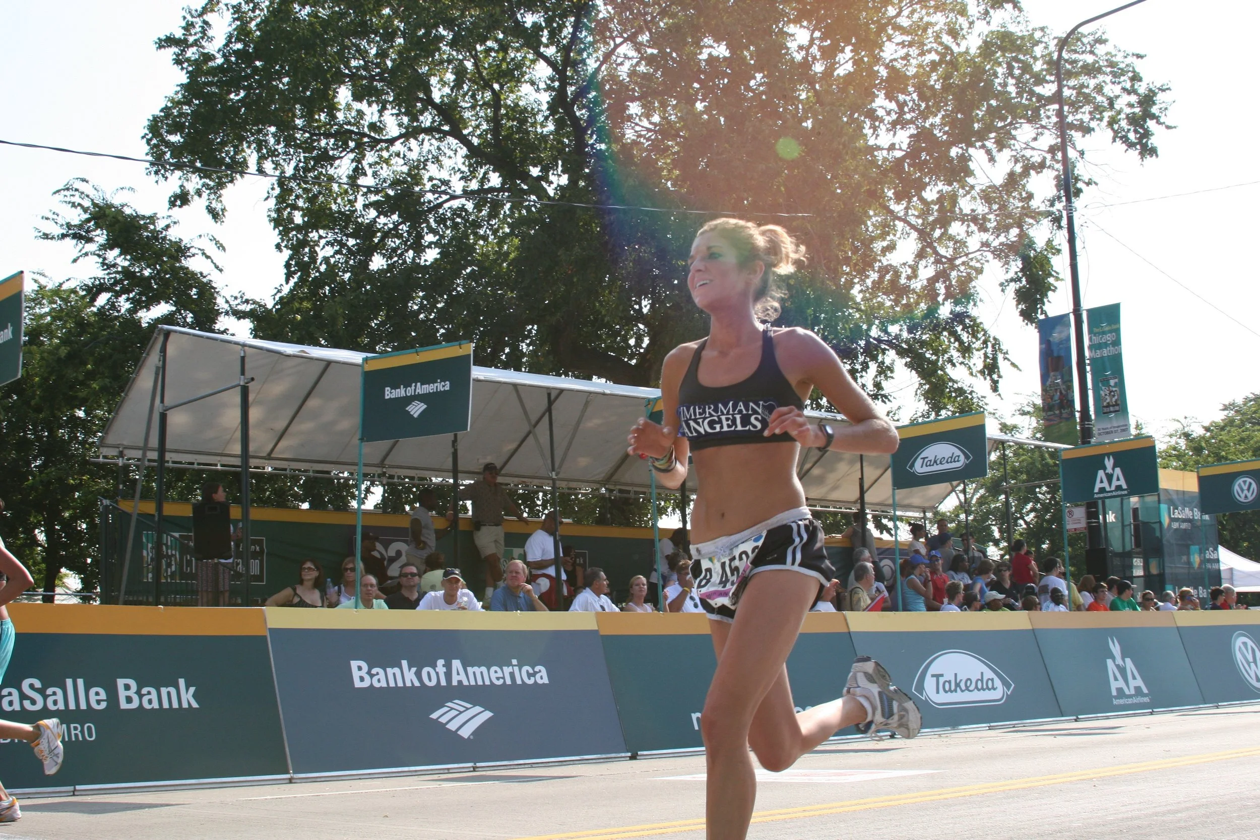 A female marathon runner in running attire is crossing the finish line at a race, with a crowd of spectators behind her and a stage with sponsorship banners in the background.