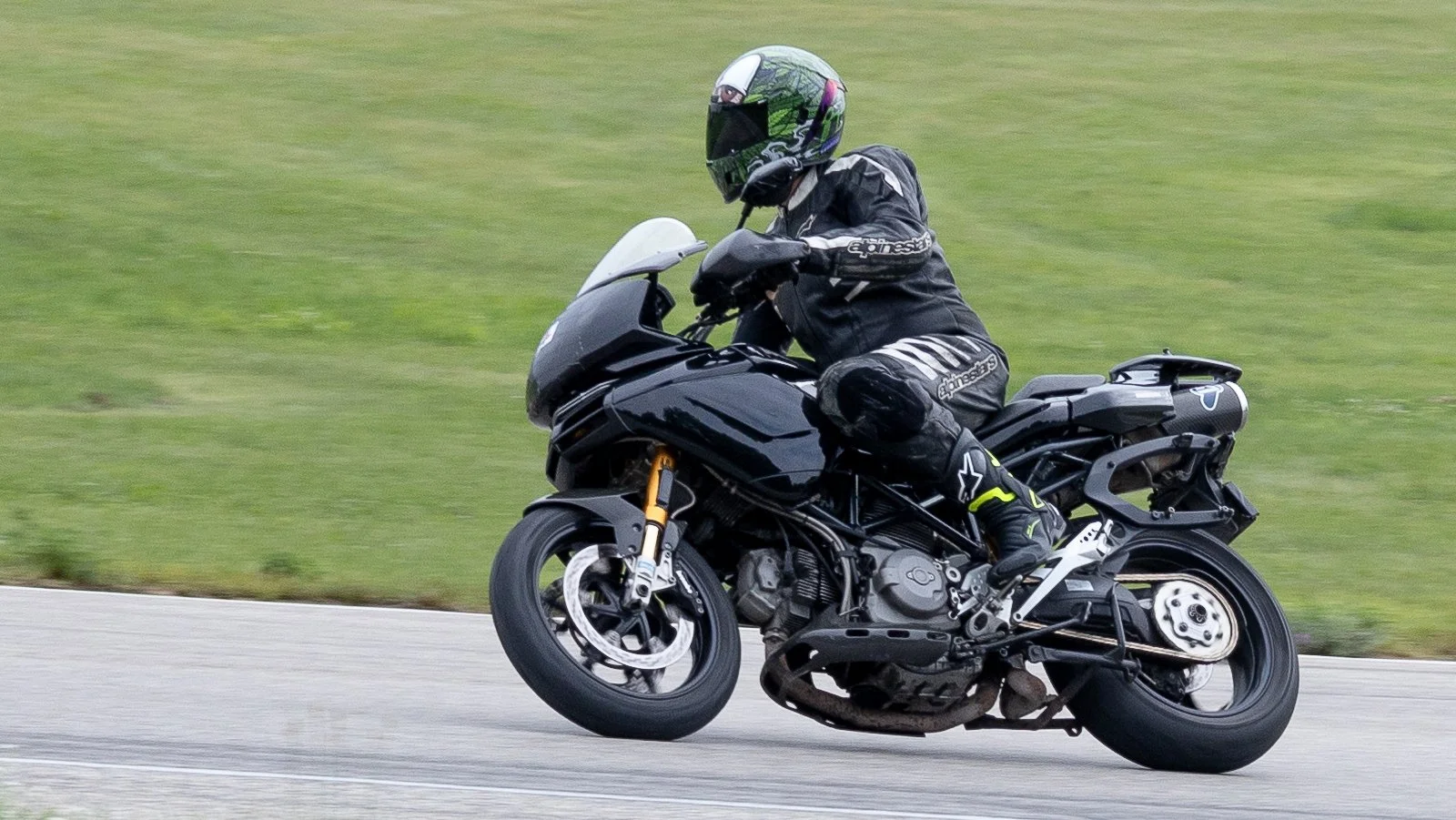 Motorcycle rider in black protective gear and a helmet leaning into a turn on a black sportbike on a paved road with green grass in the background.
