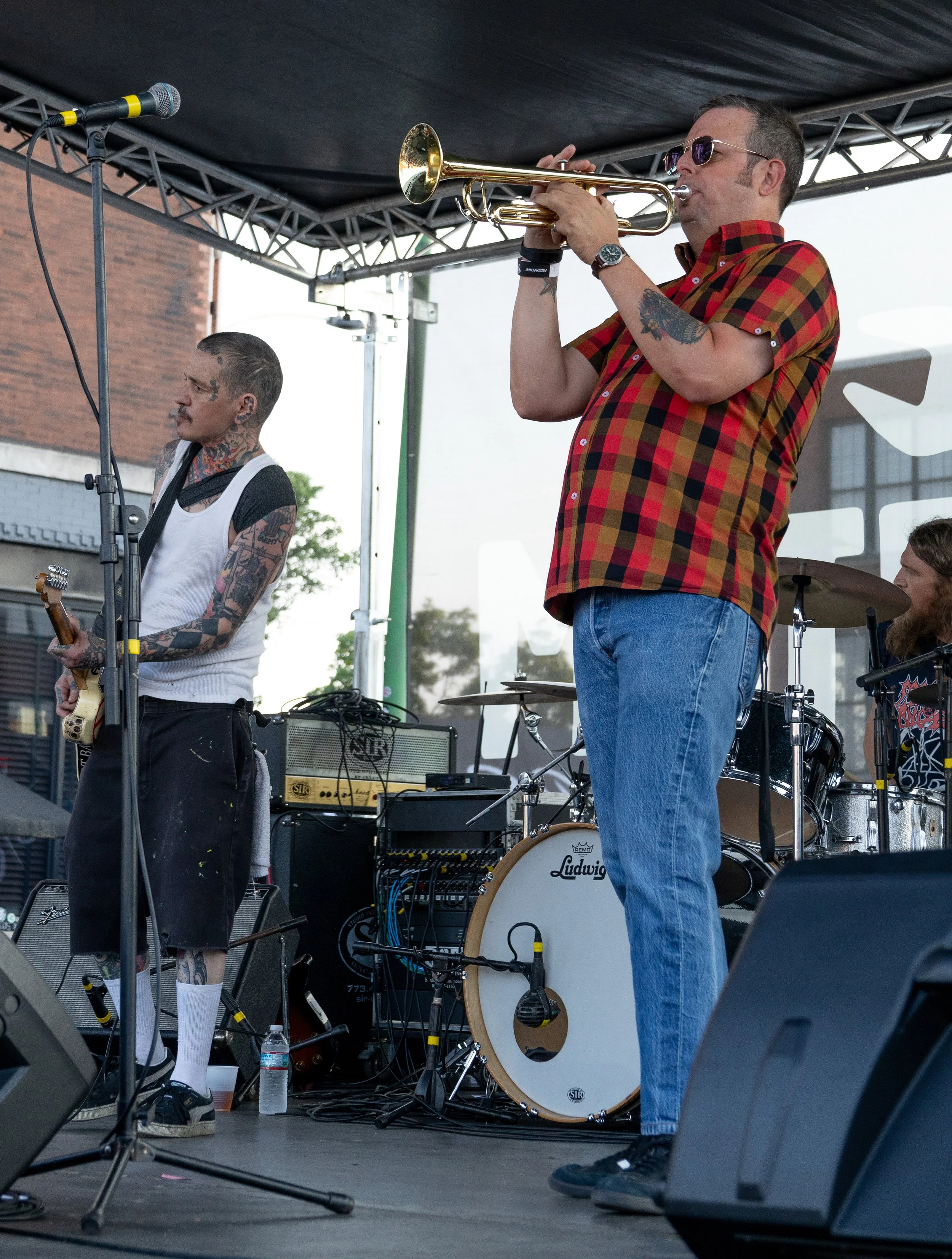 Two musicians on stage, one playing trumpet and the other playing guitar, with musical equipment including drums and amplifiers in the background during an outdoor performance.
