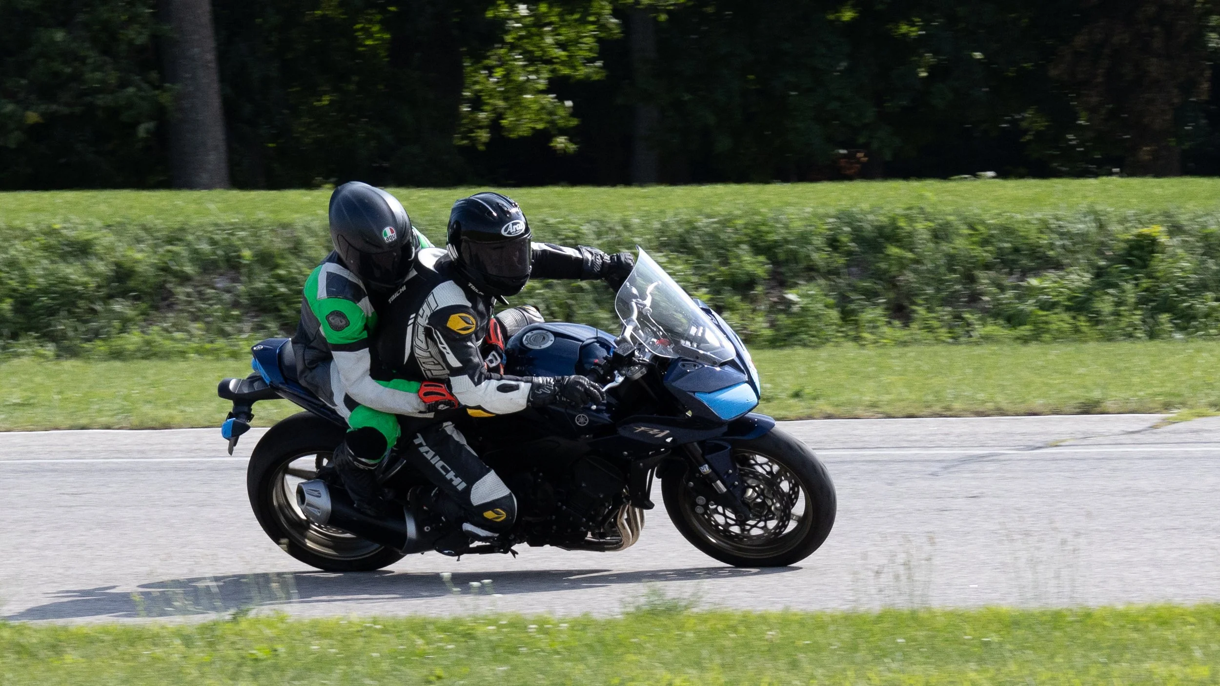 A motorcyclist wearing black helmet and racing gear riding a black Yamaha motorcycle on a road with green grass and trees in the background.