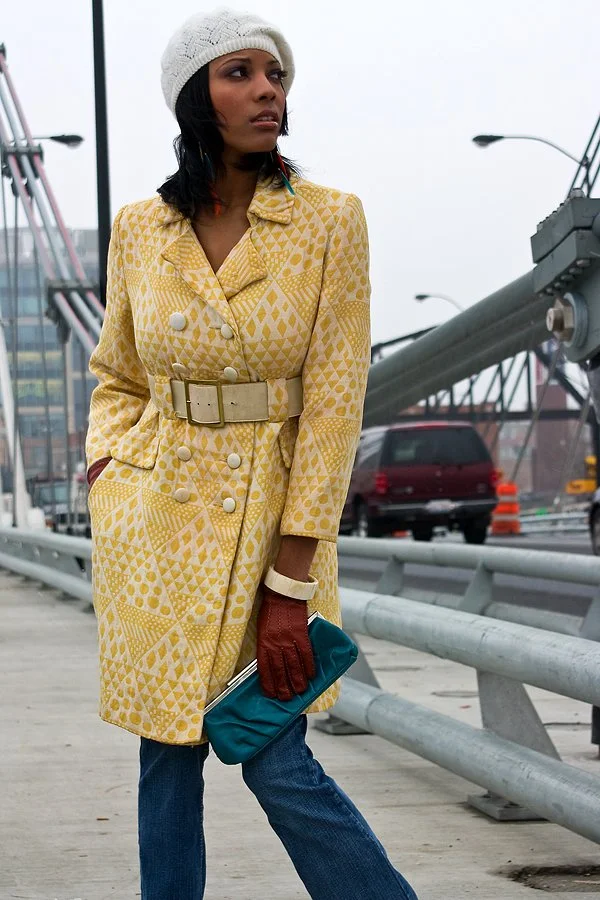 Woman in a yellow patterned coat, white knit hat, holding a teal clutch, and wearing brown gloves, standing on a bridge or sidewalk with a cityscape and parked cars in the background.