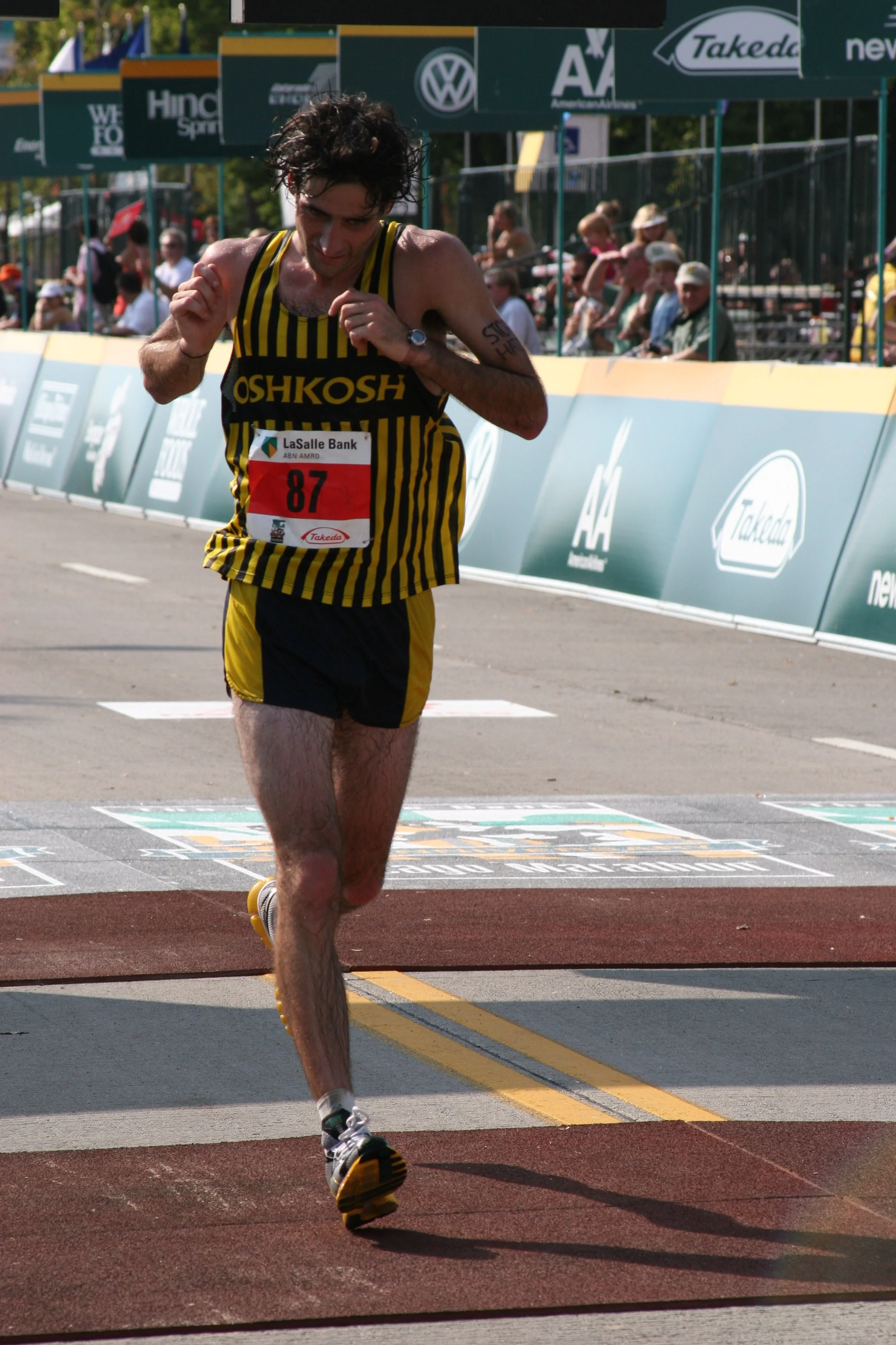 A male marathon runner in a black and yellow striped uniform nearing the finish line, with a crowd watching behind barricades and sponsorship banners along the racecourse.