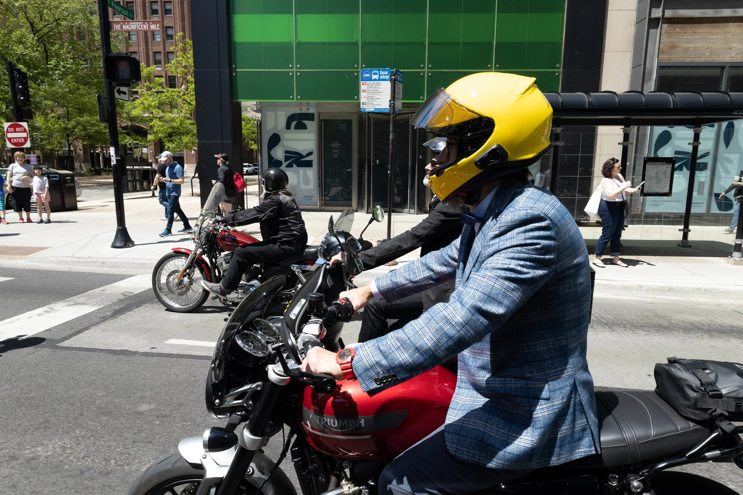 A man in a plaid jacket wearing a yellow helmet riding a red motorcycle across a city street with pedestrians and a bus stop in the background.