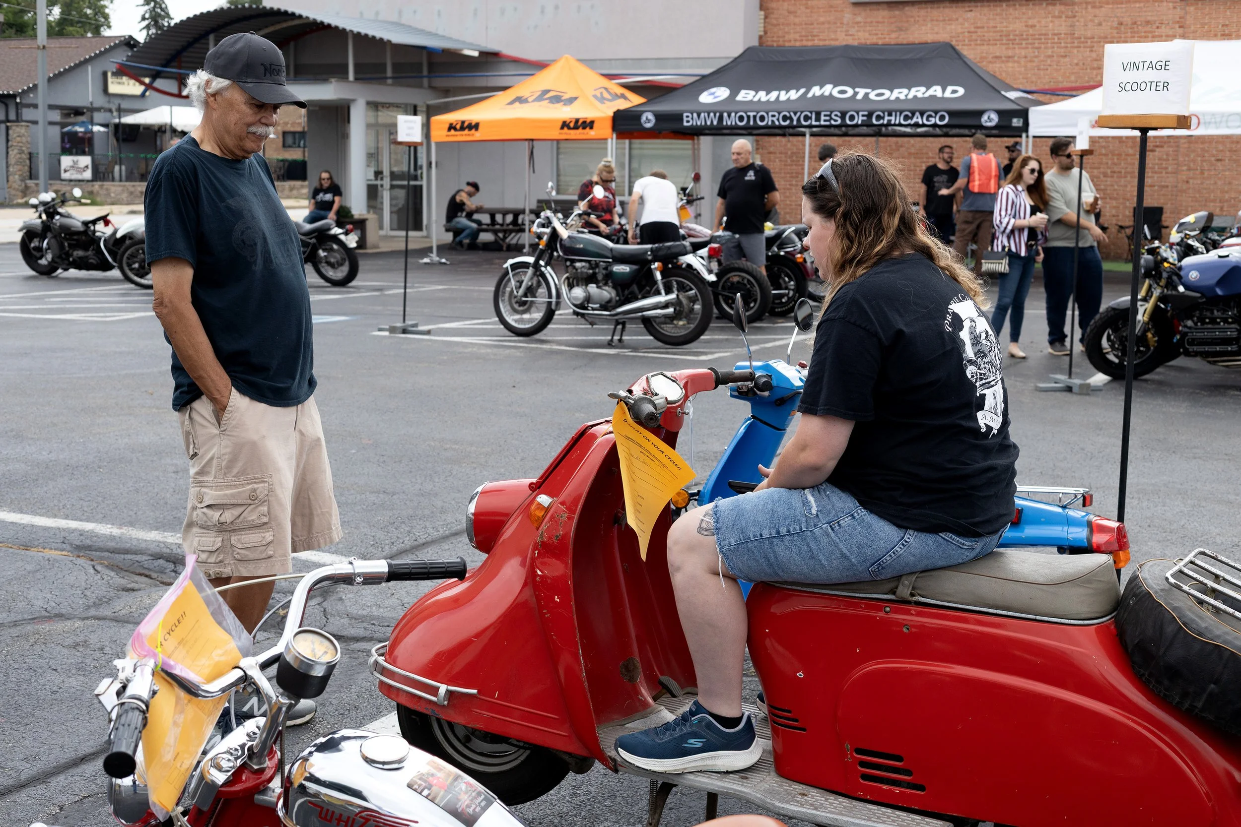 A woman in denim shorts and a black t-shirt sitting on a red scooter at a motorcycle event, talking to an older man in a navy t-shirt and beige shorts, with vintage motorcycles displayed in the background.