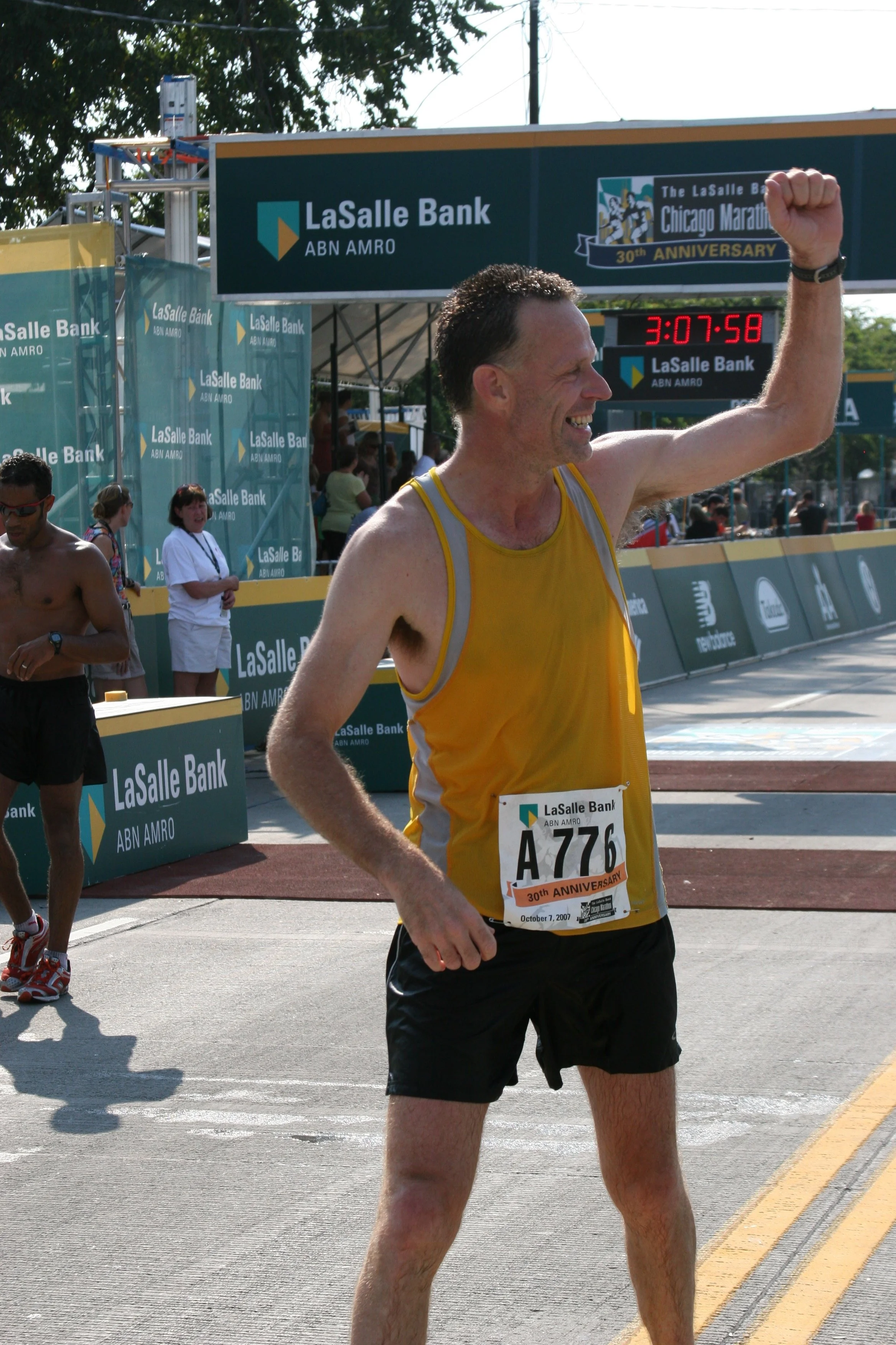 A male marathon runner in a yellow tank top and black shorts crossing the finish line, smiling with his right fist raised in victory, at the Chicago Marathon with a digital clock showing 3:07:58 and banners for LaSalle Bank and ABN AMRO in the backgr