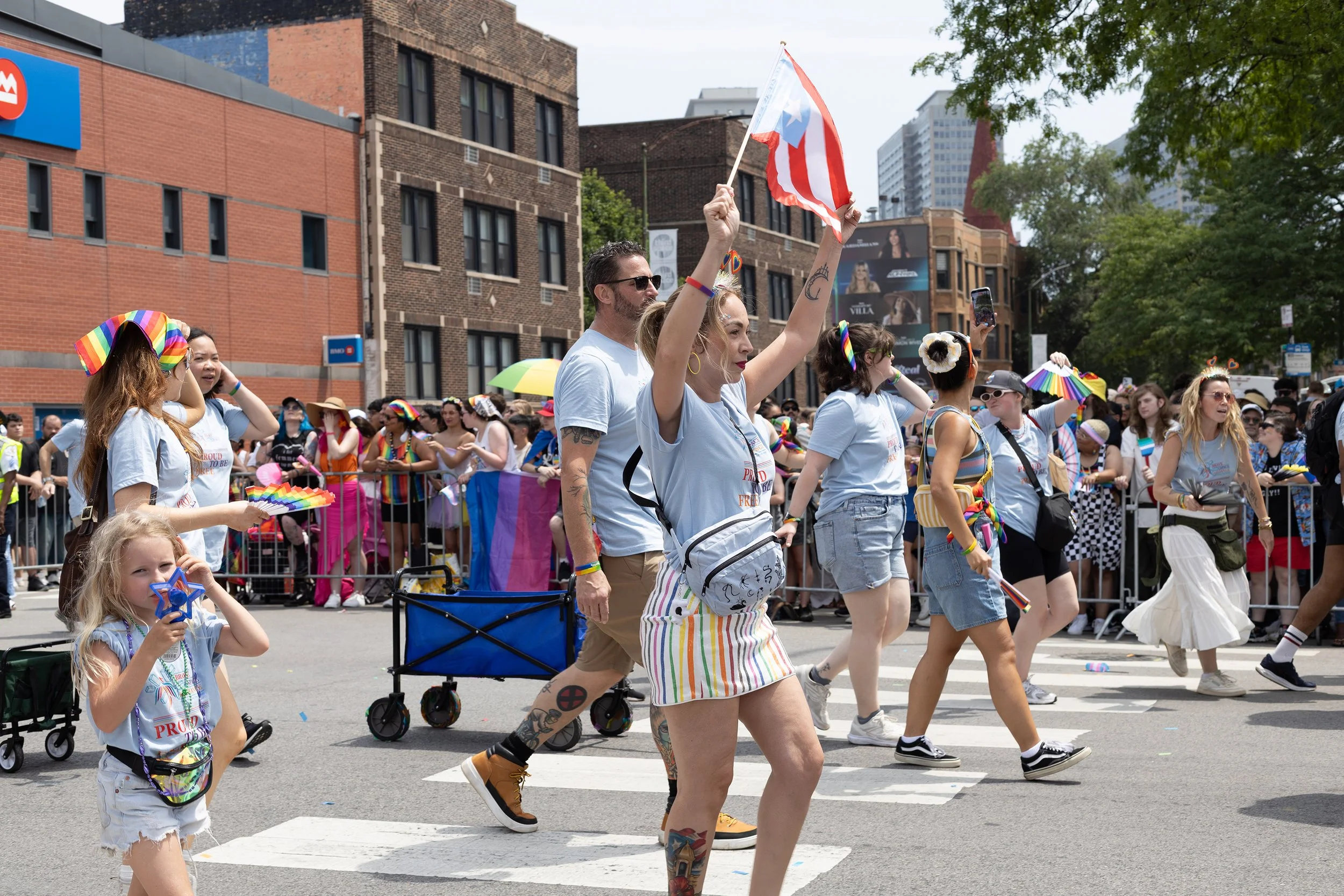 People celebrating Pride parade on city street, waving rainbow flags, wearing colorful accessories, and walking in a line with trees and buildings in the background.
