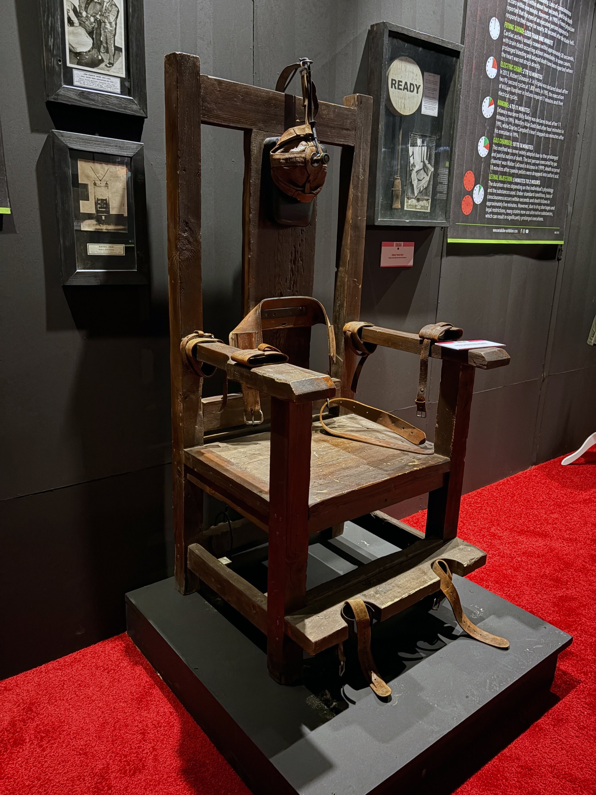 A vintage electric chair with wooden frame and straps on a display platform, with informational posters on the wall behind it.