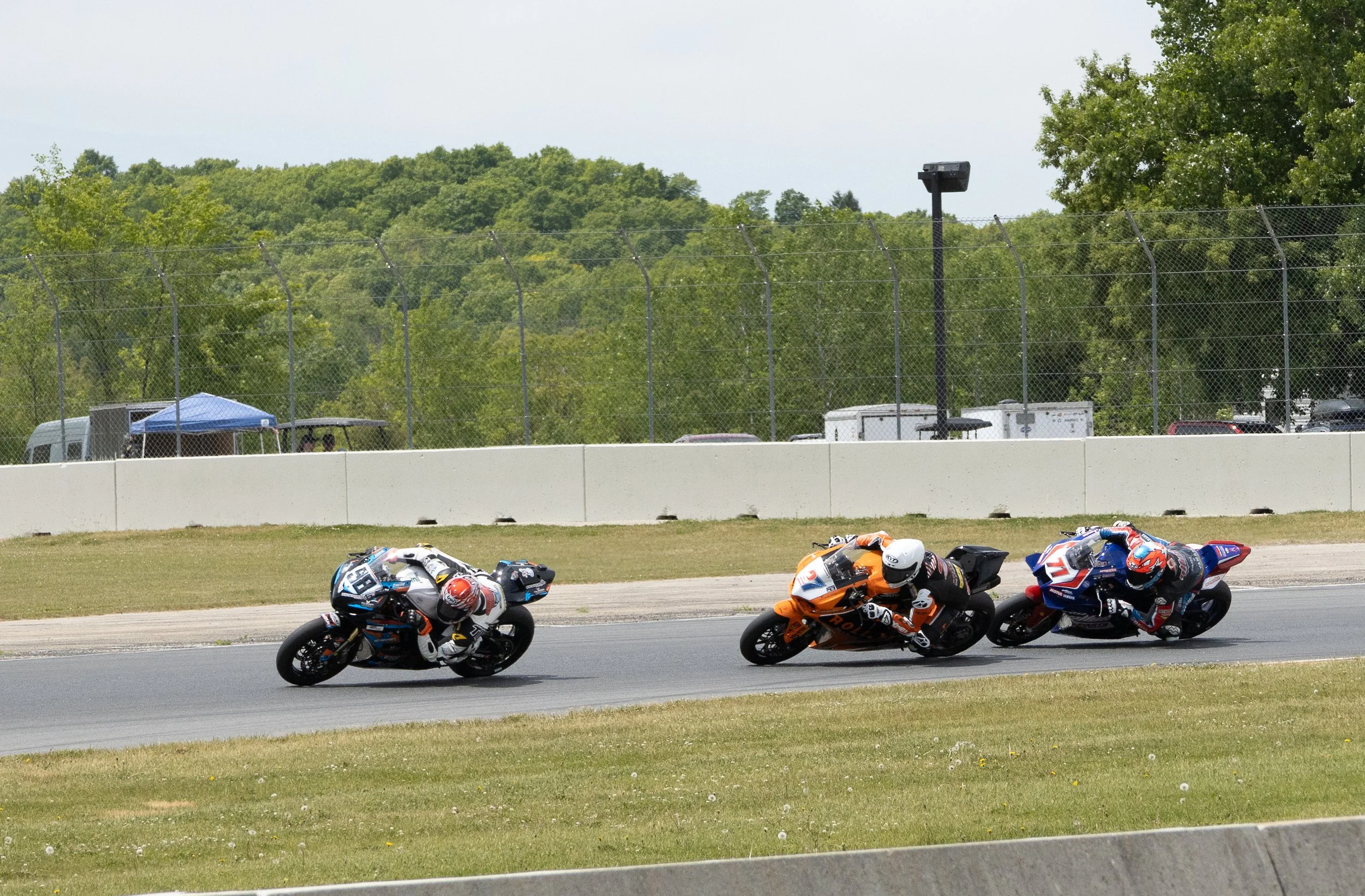 Motorcycle race on a racetrack with three riders leaning into a turn, green trees and a chain-link fence in the background.