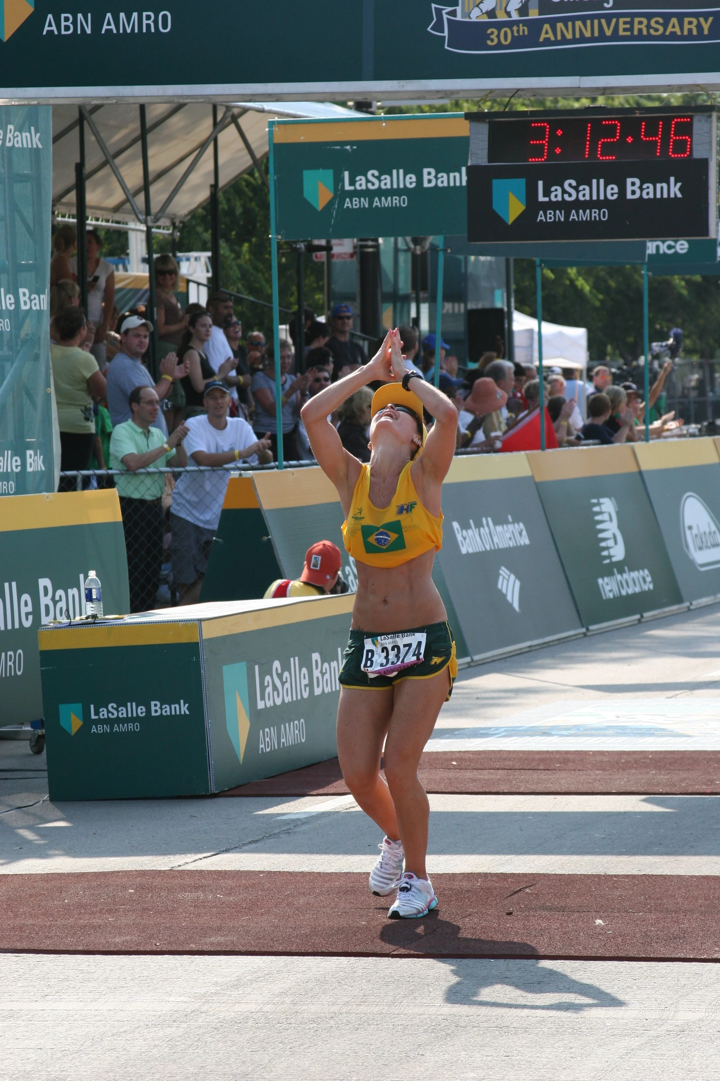 A female athlete wearing a yellow and green sports uniform with Brazil's flag logo crosses the finish line of a race, raising her hands in celebration. Spectators watch and cheer from stands behind a barrier displaying 'LaSalle Bank' and 'ABN AMRO' b