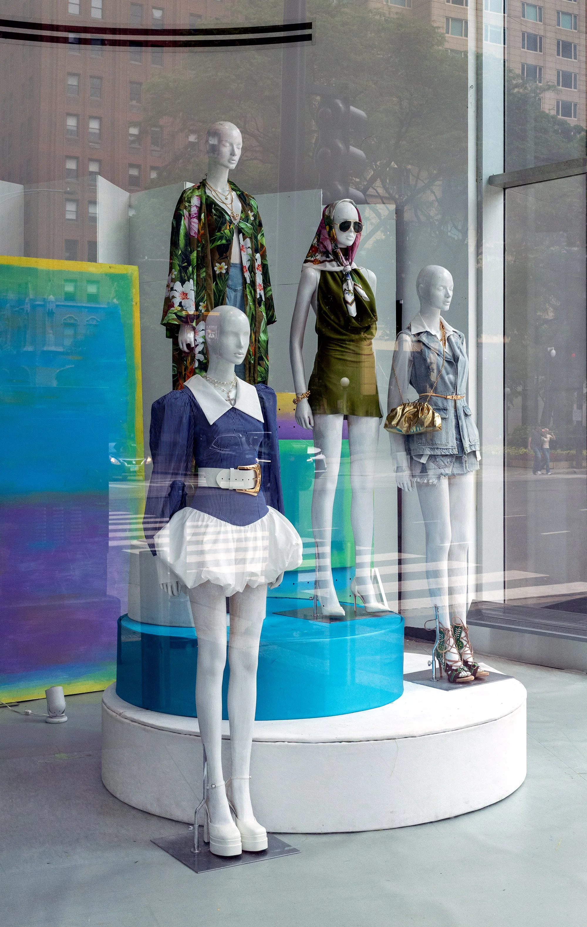 Display window featuring four mannequin torsos dressed in stylish clothing and accessories, with a rainbow-colored backdrop and city buildings reflected in the glass.