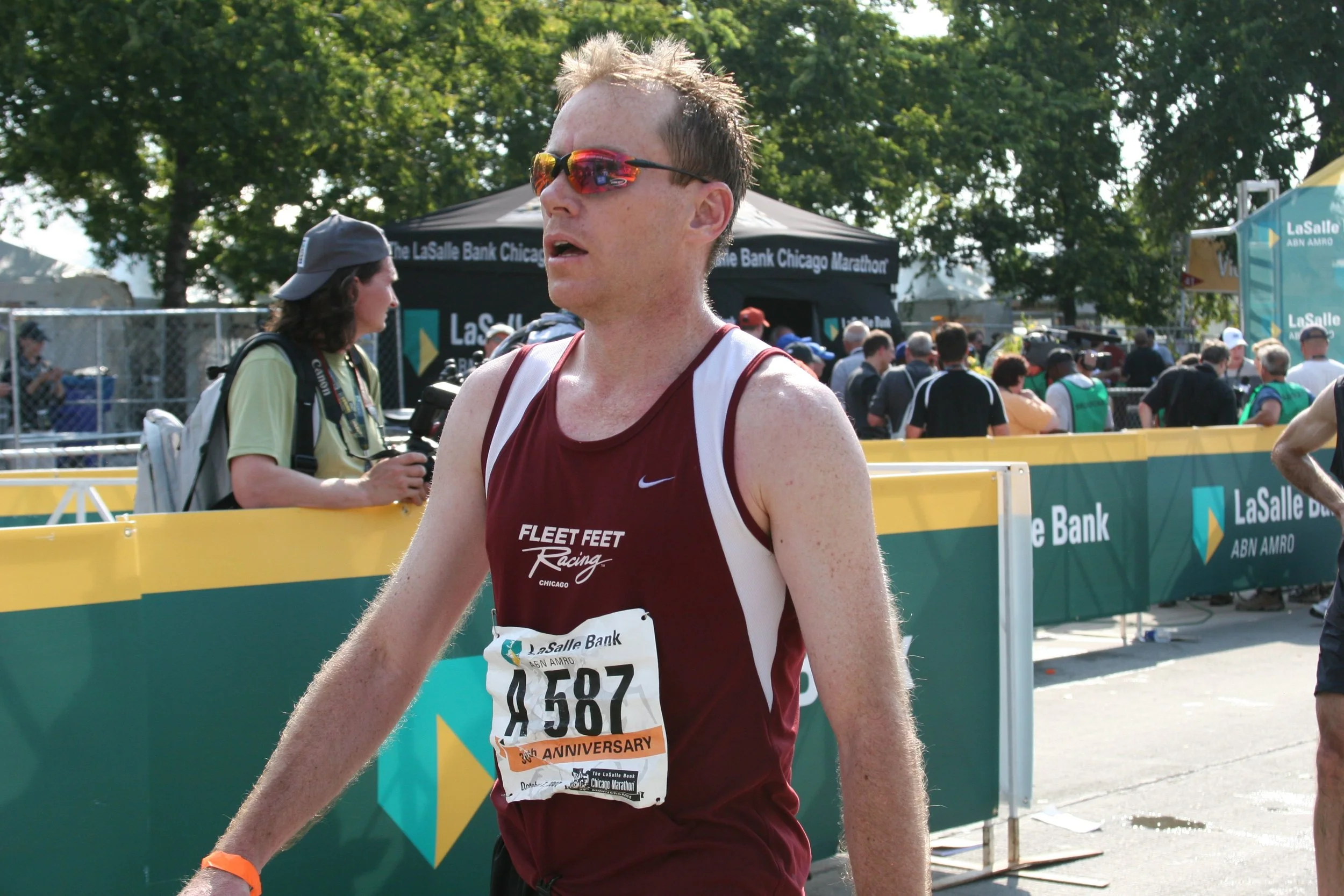 A male marathon runner wearing red sunglasses, a maroon and white tank top, and a race bib, crossing the finish line at the Chicago Marathon with trees and spectators in the background.