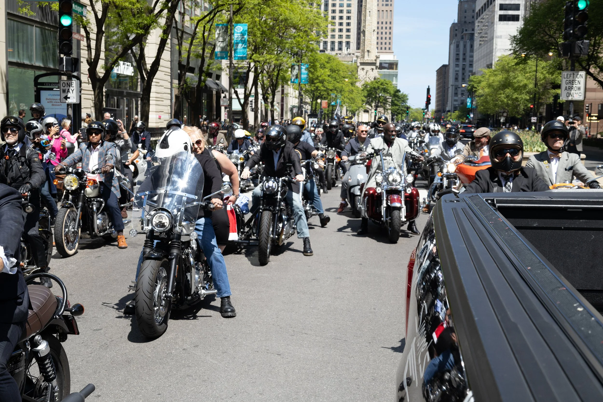A large crowd of people on motorcycles and scooters waiting at a city street intersection with tall buildings and green trees, during a sunny day.