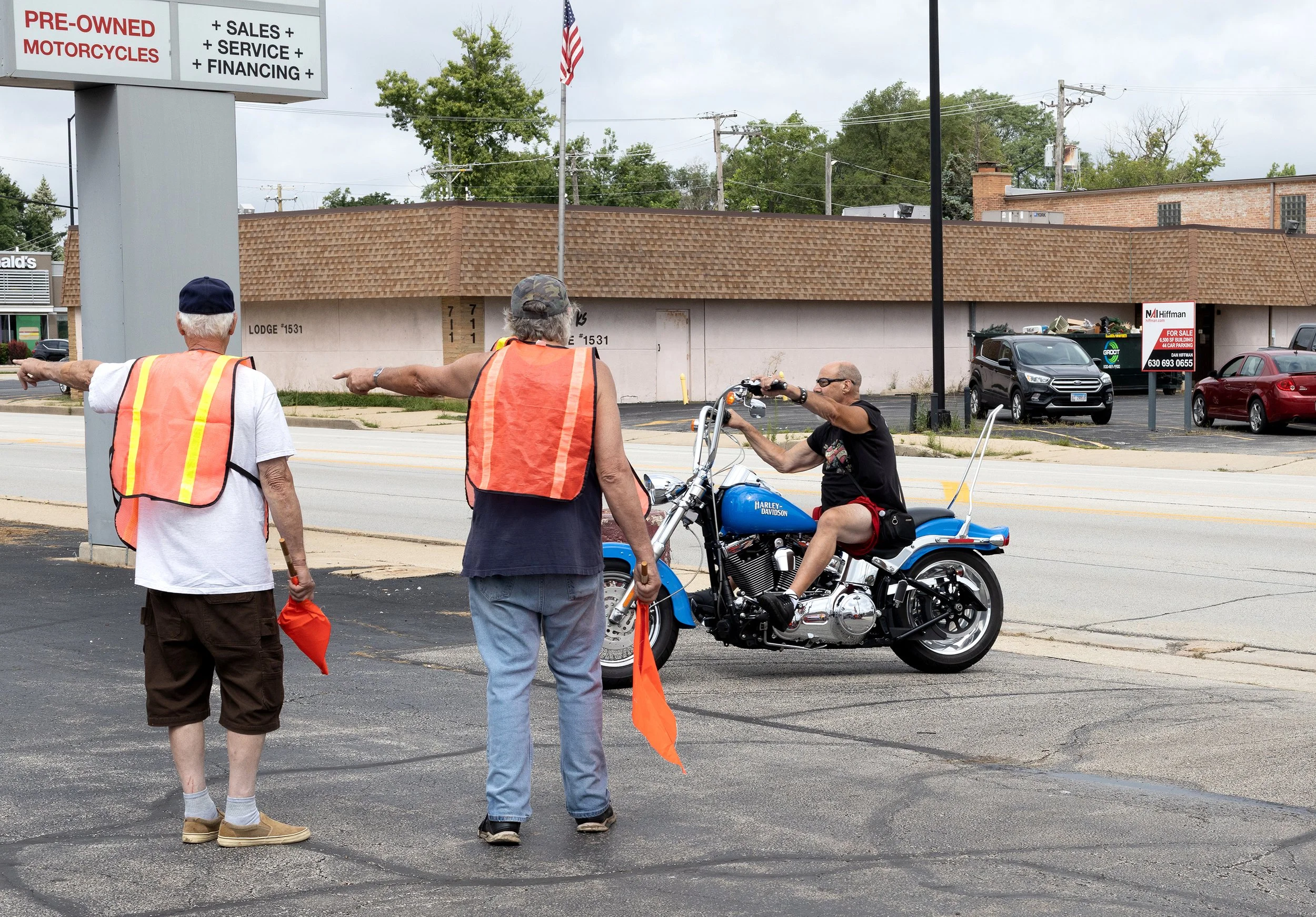 Three people, two women and one man, wearing orange safety vests, are standing on the sidewalk. The man is sitting on a blue Harley-Davidson motorcycle, and the women are pointing across the street, possibly directing or signaling. In the background,