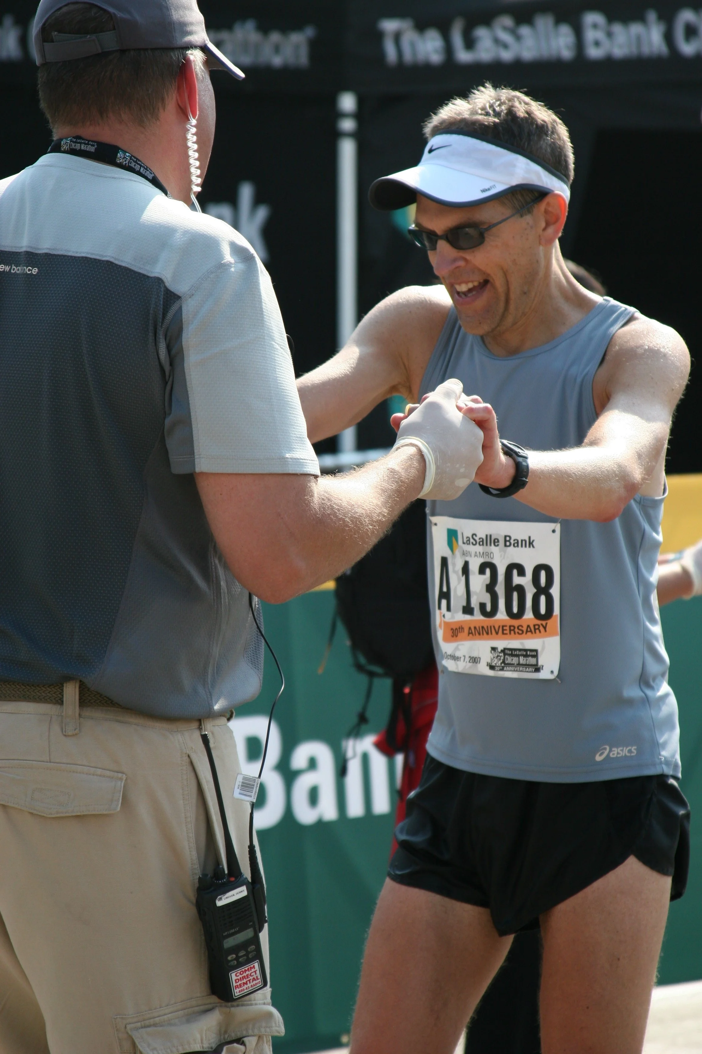 A male marathon runner in athletic gear, wearing a visor and sunglasses, is smiling and exchanging a handshake with an official or volunteer, who is wearing a gray shirt and beige pants. The runner has a race bib with number A1368 and the word 'Anniv