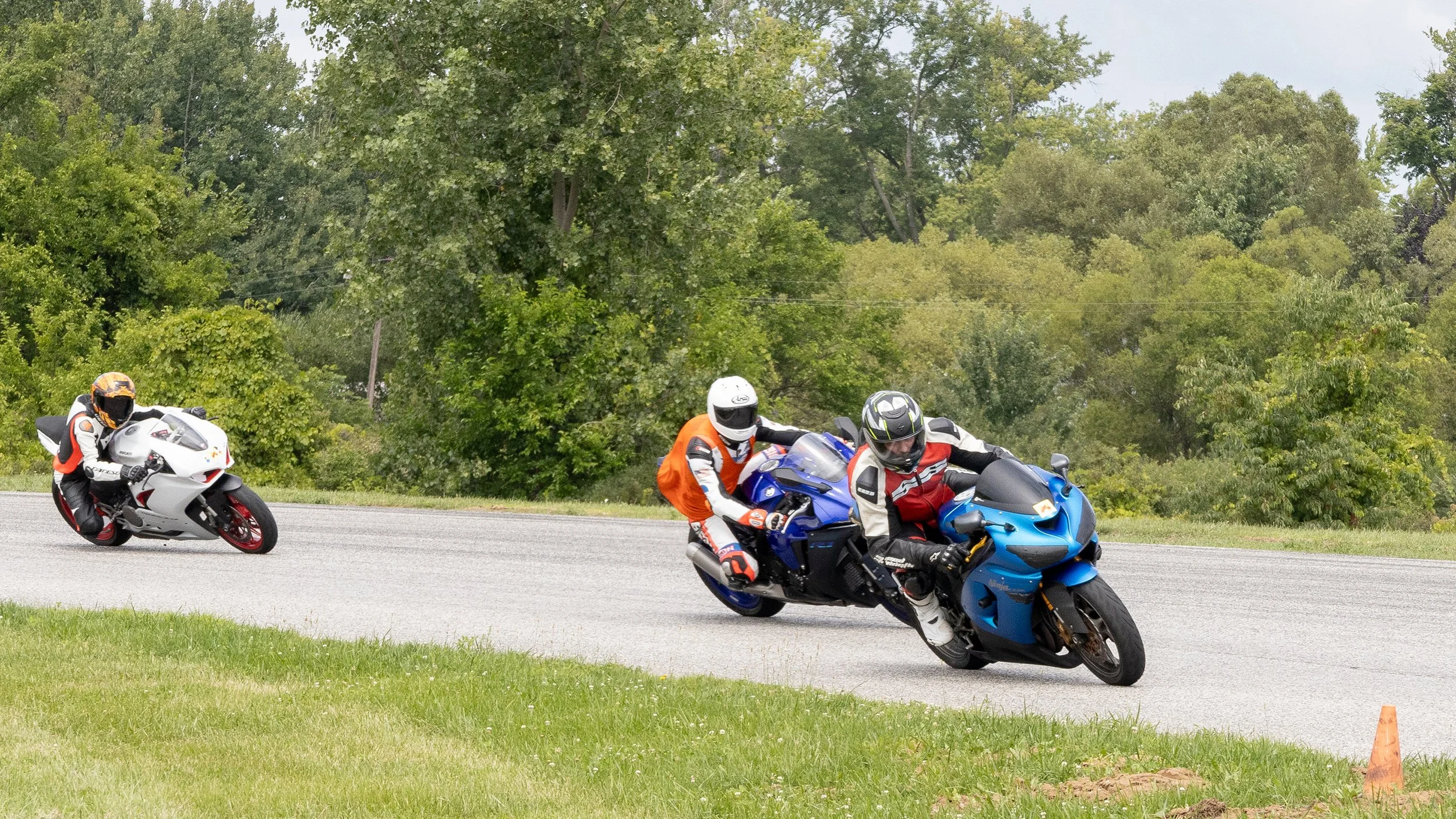 Three motorcyclists riding on a paved road surrounded by green trees. They are wearing protective gear, including helmets and racing suits, leaning into a turn as they navigate the curve.