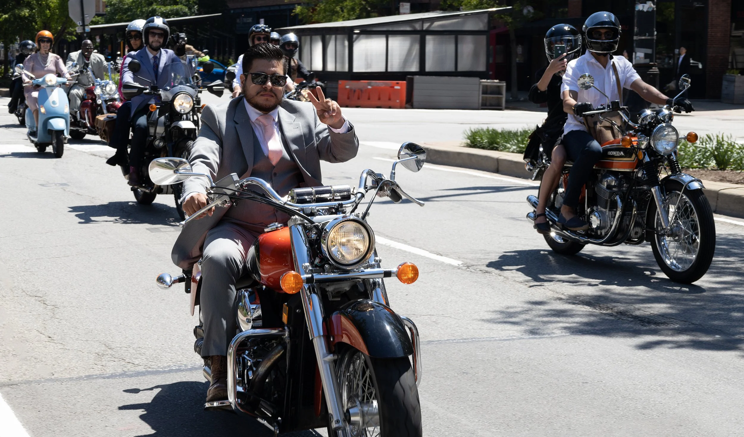 A group of people riding motorcycles on a city street, with a man in the front making a peace sign and wearing sunglasses, and others behind him also wearing helmets.