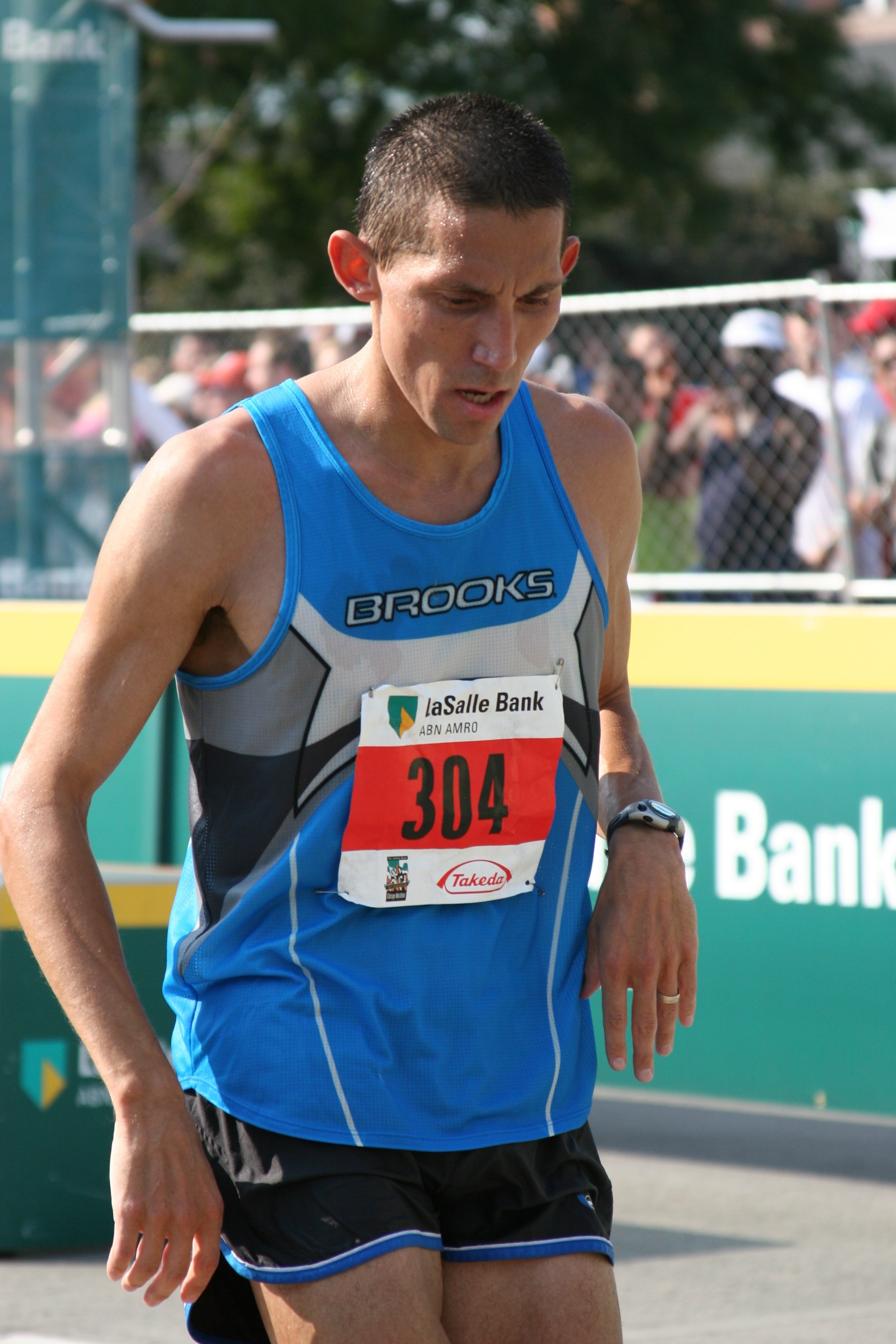 Male runner in blue athletic gear with race bib number 304, at a sporting event, with spectators behind a fence.