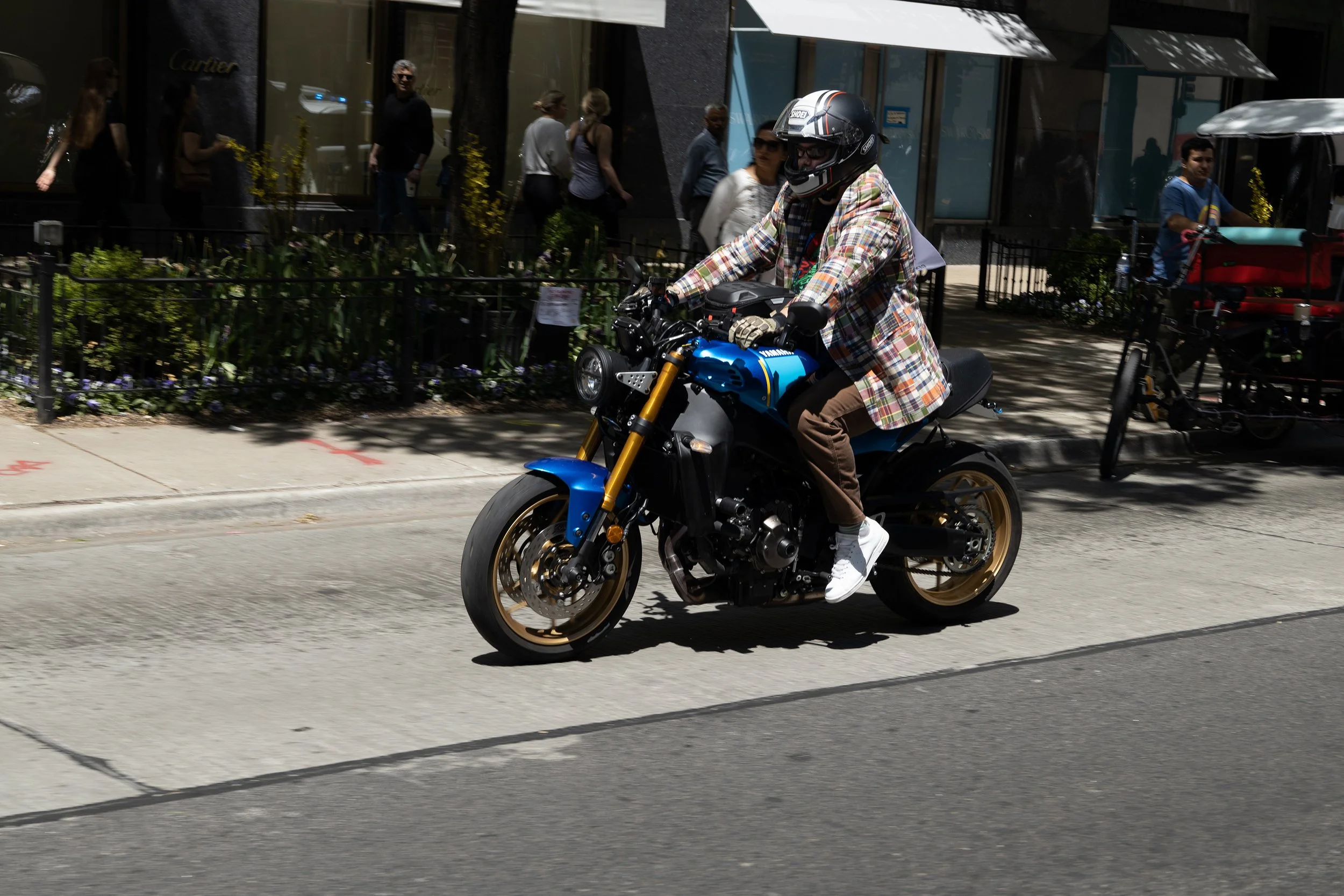 A person wearing a plaid shirt, brown pants, white shoes, and a motorcycle helmet riding a blue Yamaha motorcycle on a city street with pedestrians and storefronts in the background.
