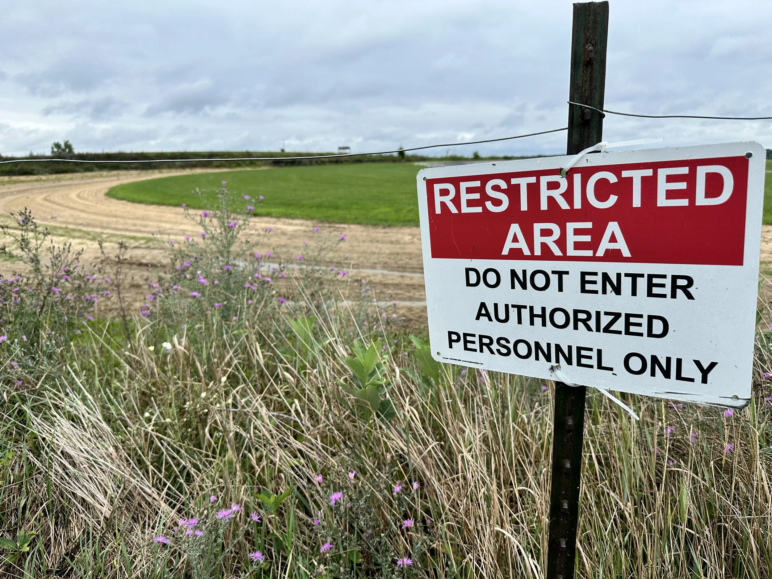 Sign that reads 'Restricted Area Do Not Enter Authorized Personnel Only' located in a rural area with a dirt path, green grass, and purple wildflowers, under a cloudy sky.