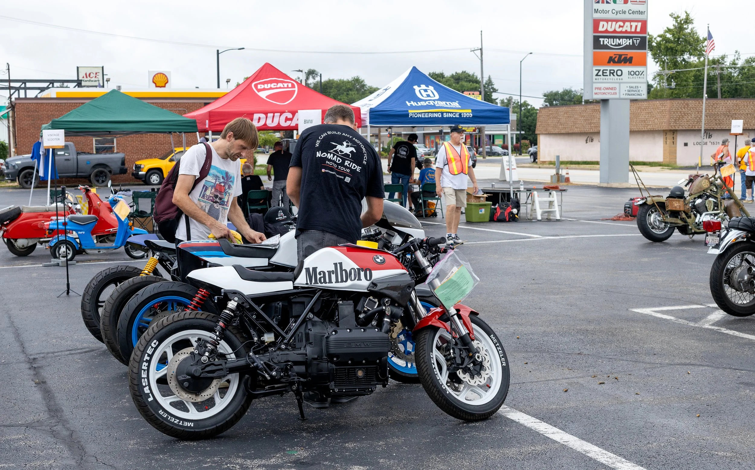 Multiple motorcycles displayed in a parking lot during an outdoor event with tents and people inspecting the bikes.