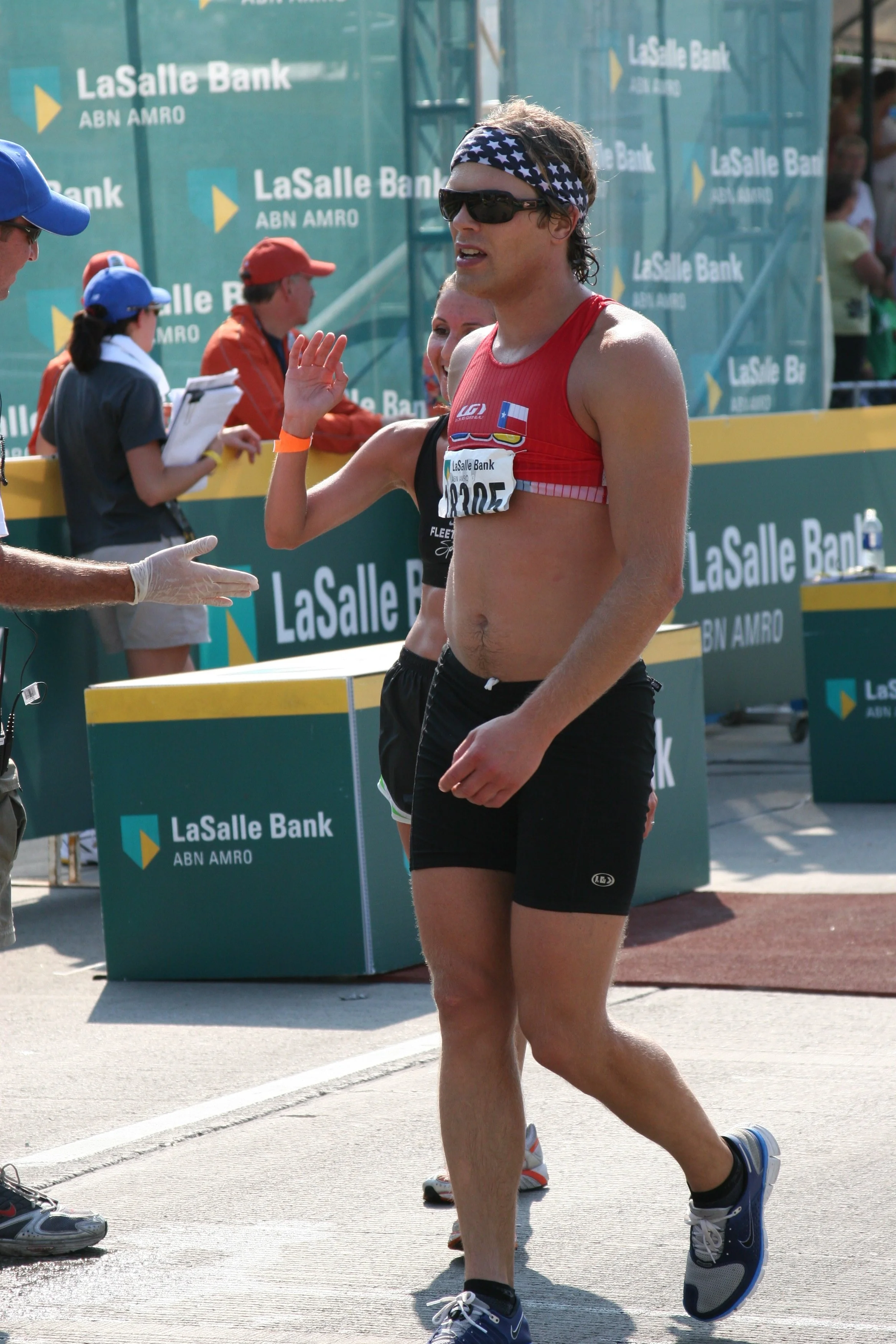 A male marathon runner in a red sleeveless shirt and black shorts running on a street, wearing sunglasses and a star-spangled headband, with a medal around his neck. There are race officials and fans on the side, and signs for LaSalle Bank and ABN AM