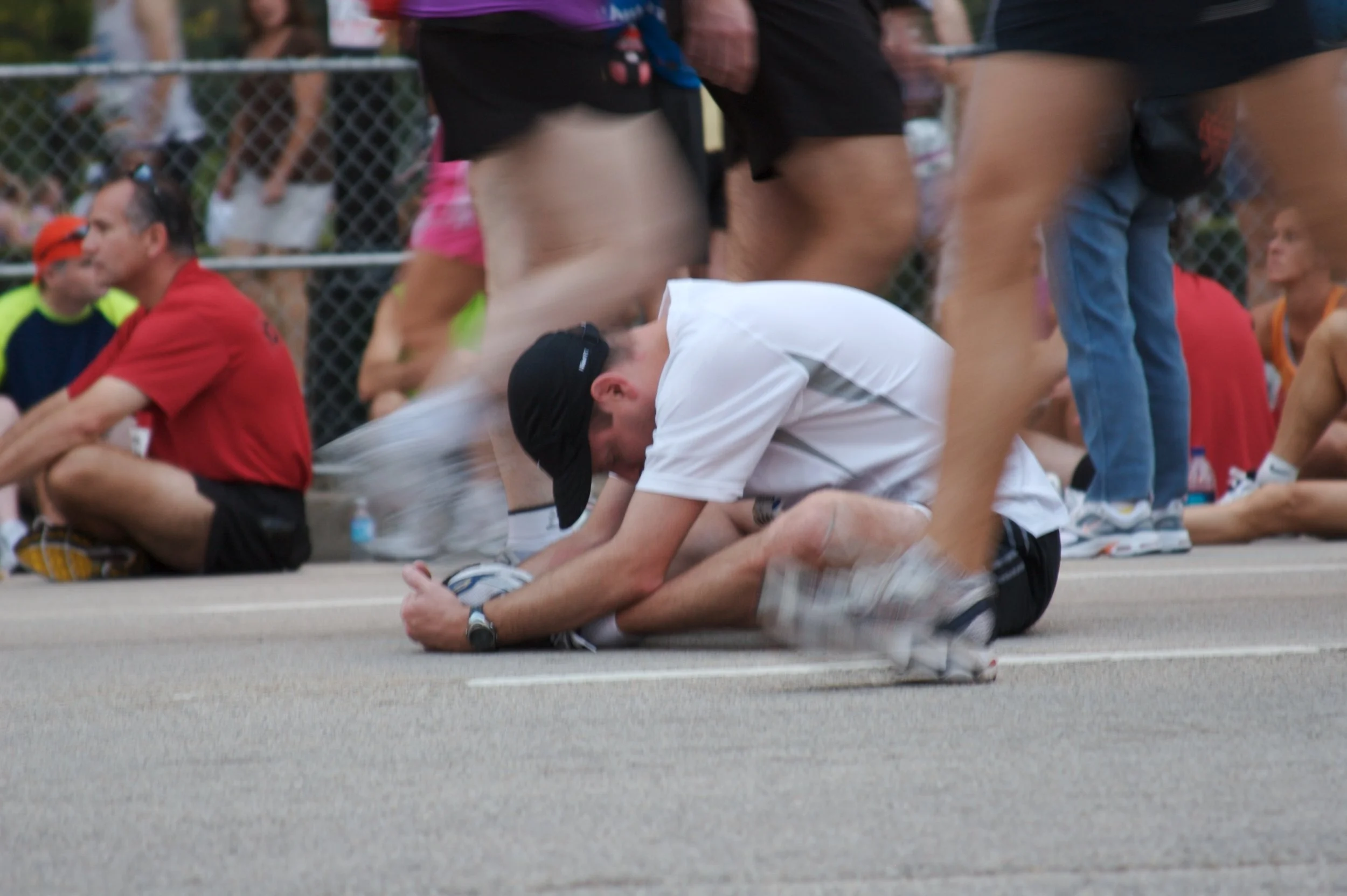 A person kneeling on the ground during a marathon, surrounded by spectators and other runners, some sitting and some standing behind a fence.
